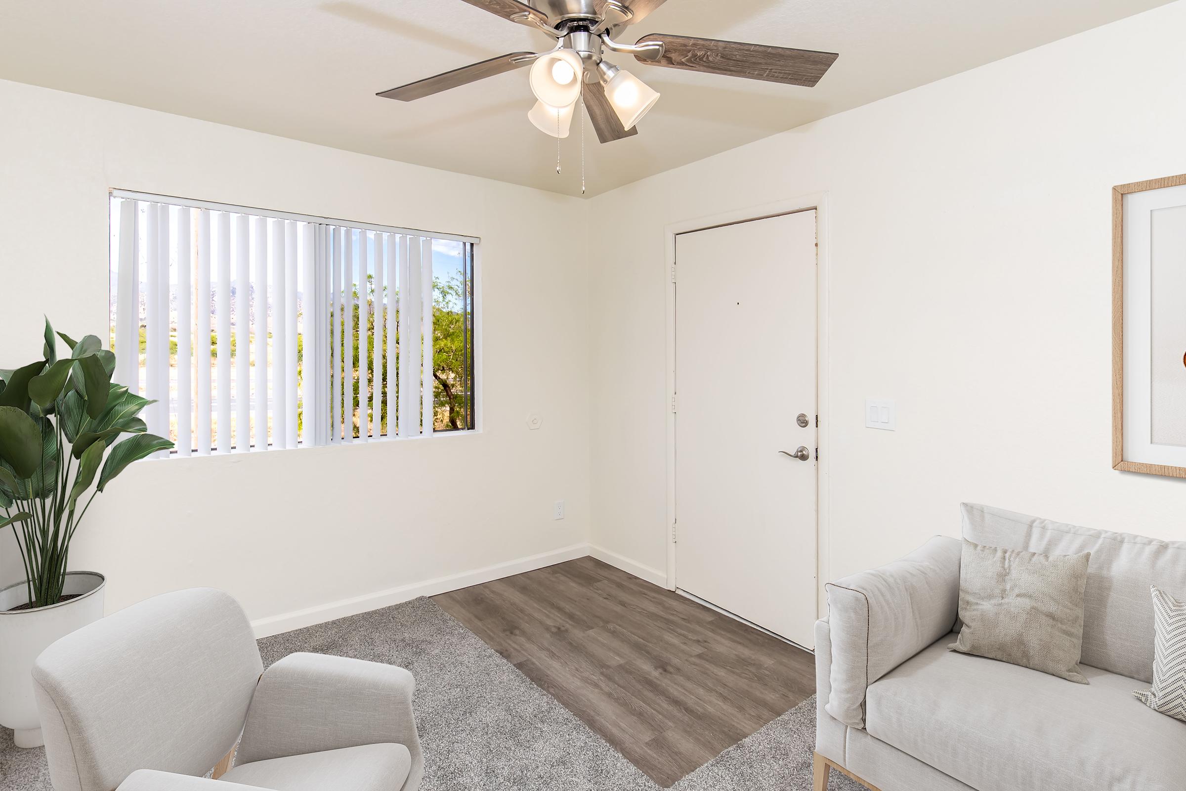 A cozy room featuring a light-colored wall, a ceiling fan with wooden blades, a window with vertical blinds, and a door. There’s a small gray sofa with cushions and a light gray armchair. A potted plant adds a touch of greenery, while soft carpeting completes the inviting atmosphere.