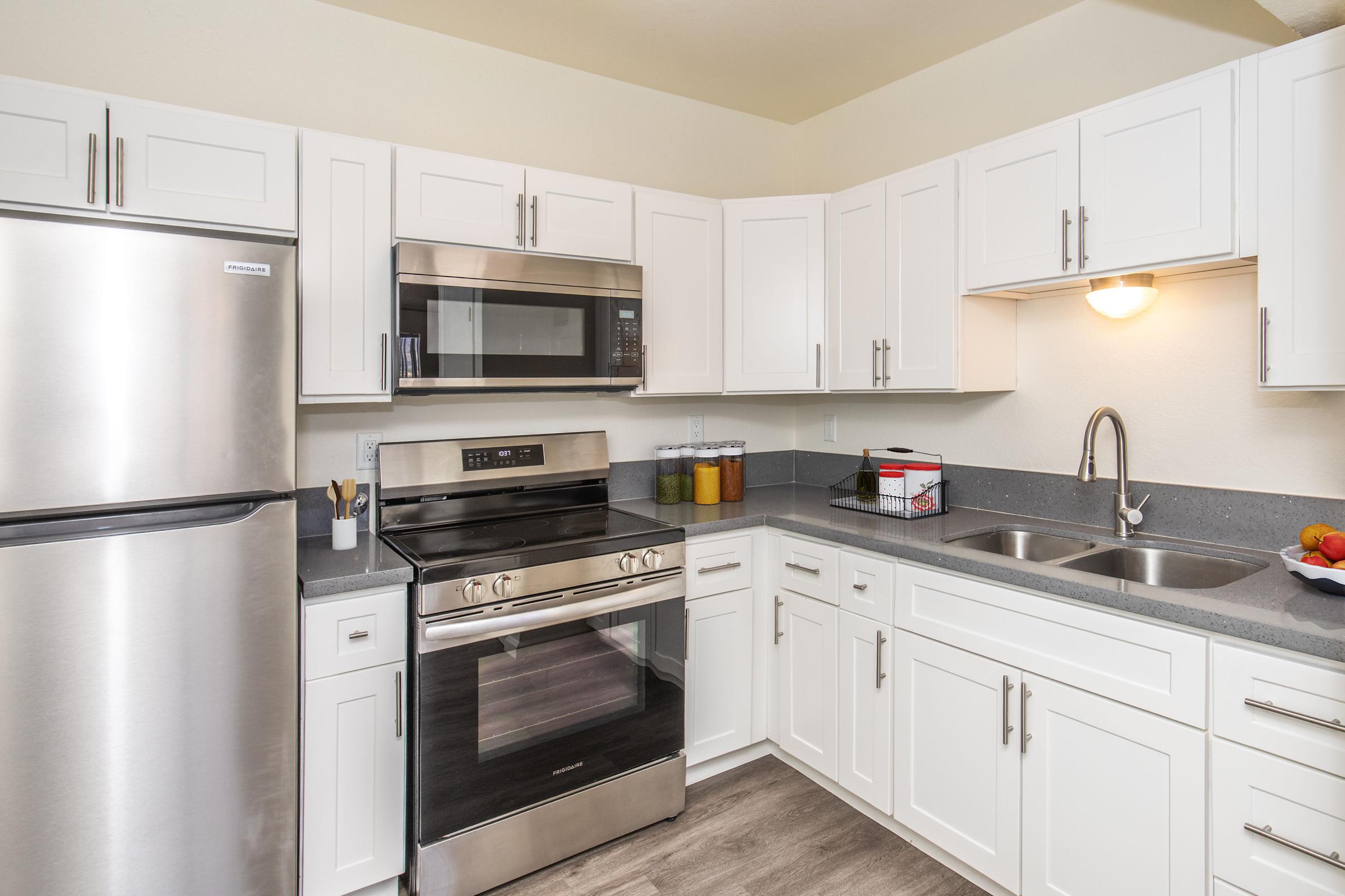 Modern kitchen featuring stainless steel appliances including a refrigerator, microwave, and oven. White cabinetry and gray countertops provide a clean, bright look. A sink is positioned near a window, with bowls of fruit on the counter, creating a welcoming and functional cooking space.
