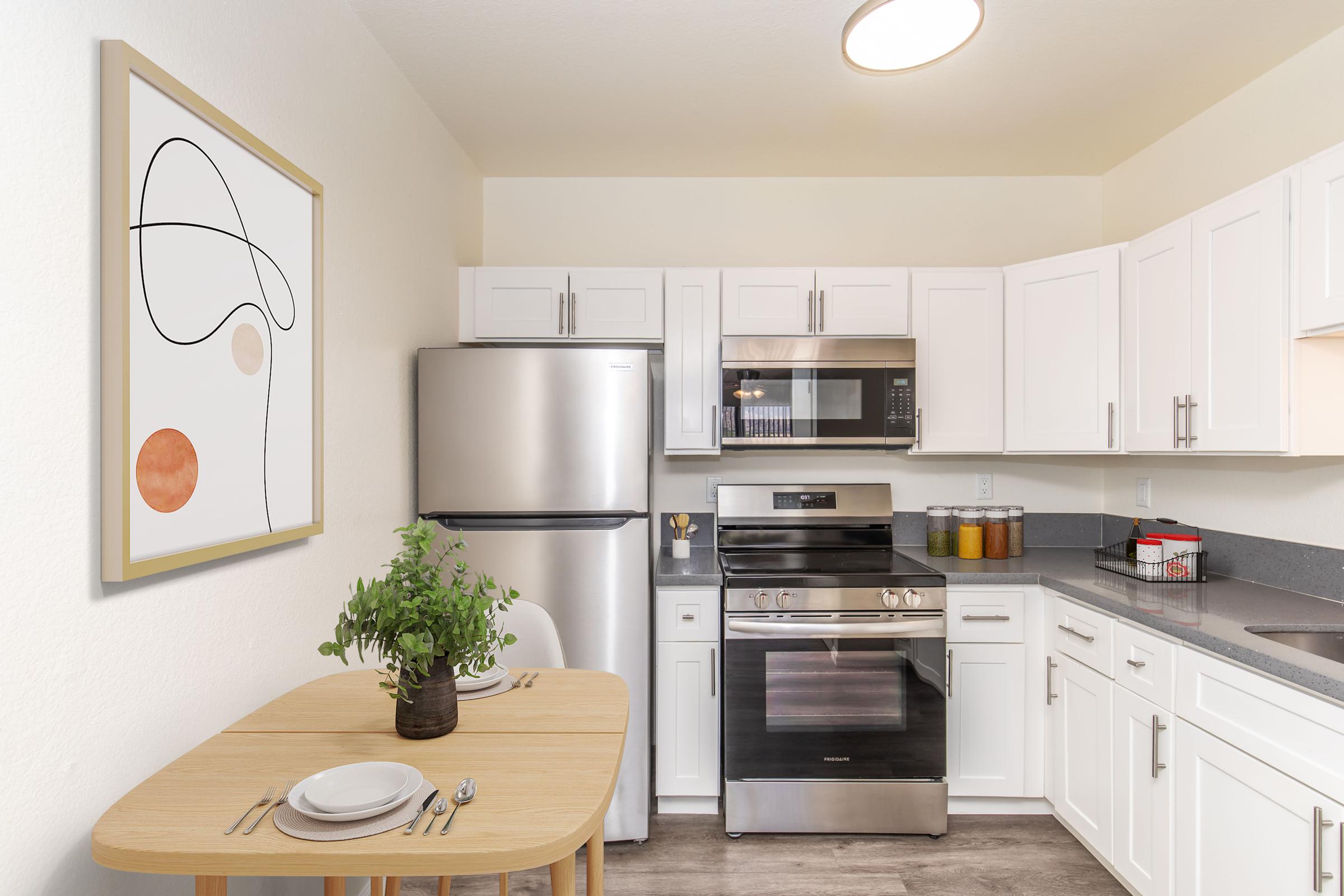 A modern kitchen featuring white cabinetry, stainless steel appliances, and a gray countertop. A small round dining table is set for two with plates and utensils. A piece of abstract art hangs on the wall, adding a touch of color to the space. Natural light enhances the clean and minimalistic design.