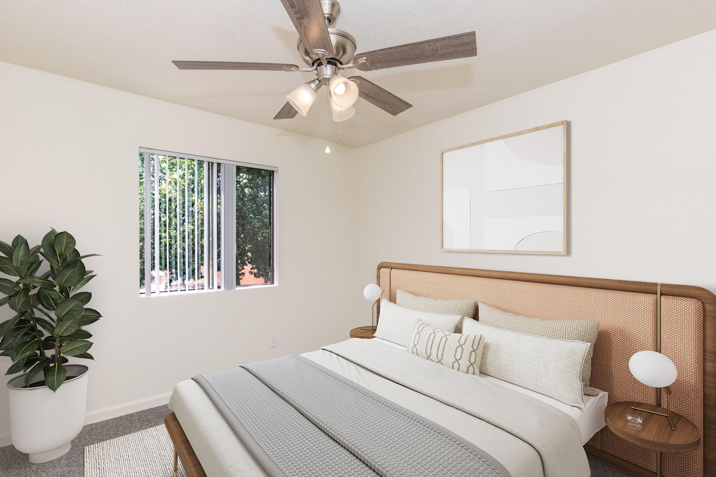 A modern bedroom featuring a king-sized bed with neutral-toned bedding, decorative pillows, and a stylish headboard. There is a ceiling fan with light fixtures, a large window with vertical blinds, and a potted plant in the corner. An abstract art piece is mounted on the wall.