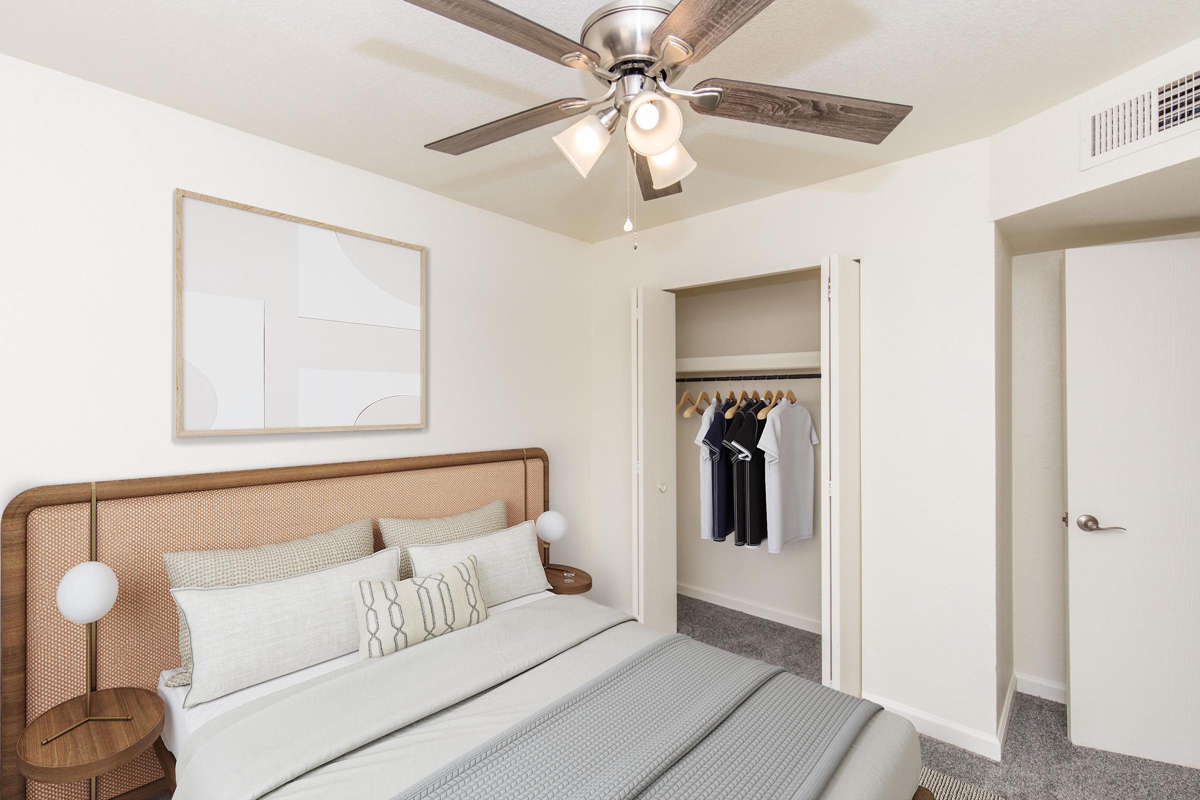 A cozy bedroom featuring a modern design with a wooden bed frame, decorative pillows, and a light gray blanket. The room includes a ceiling fan with lights, a framed abstract artwork on the wall, and an open closet revealing neatly hung clothes. Soft carpet flooring enhances the inviting atmosphere.