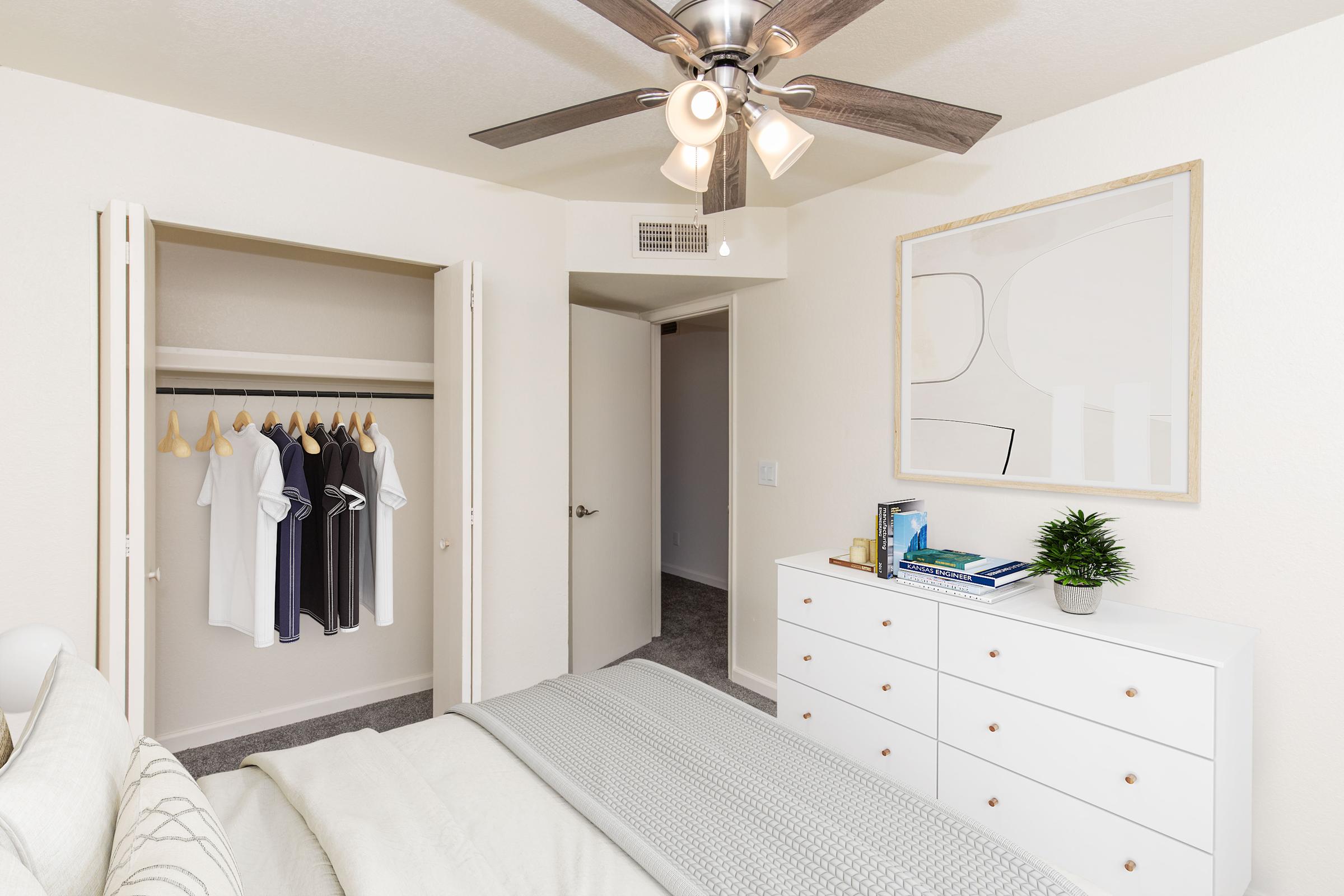 A cozy bedroom featuring a light color palette. There is a neatly made bed with a gray blanket, a modern dresser with books and a small plant, and a closet with hanging clothes. A ceiling fan adds a functional touch, and a door leads to another room, enhancing the open and inviting atmosphere.