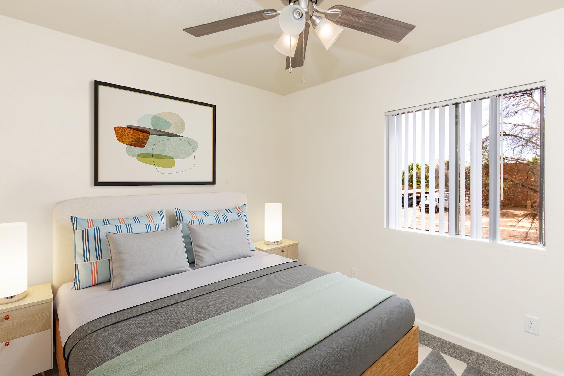 A brightly lit bedroom featuring a queen-sized bed with blue and gray bedding, bedside lamps on wooden nightstands, and a framed abstract artwork on the wall. A window with blinds provides natural light, and a view of the outside is visible. The walls are painted in light colors, creating a calm atmosphere.