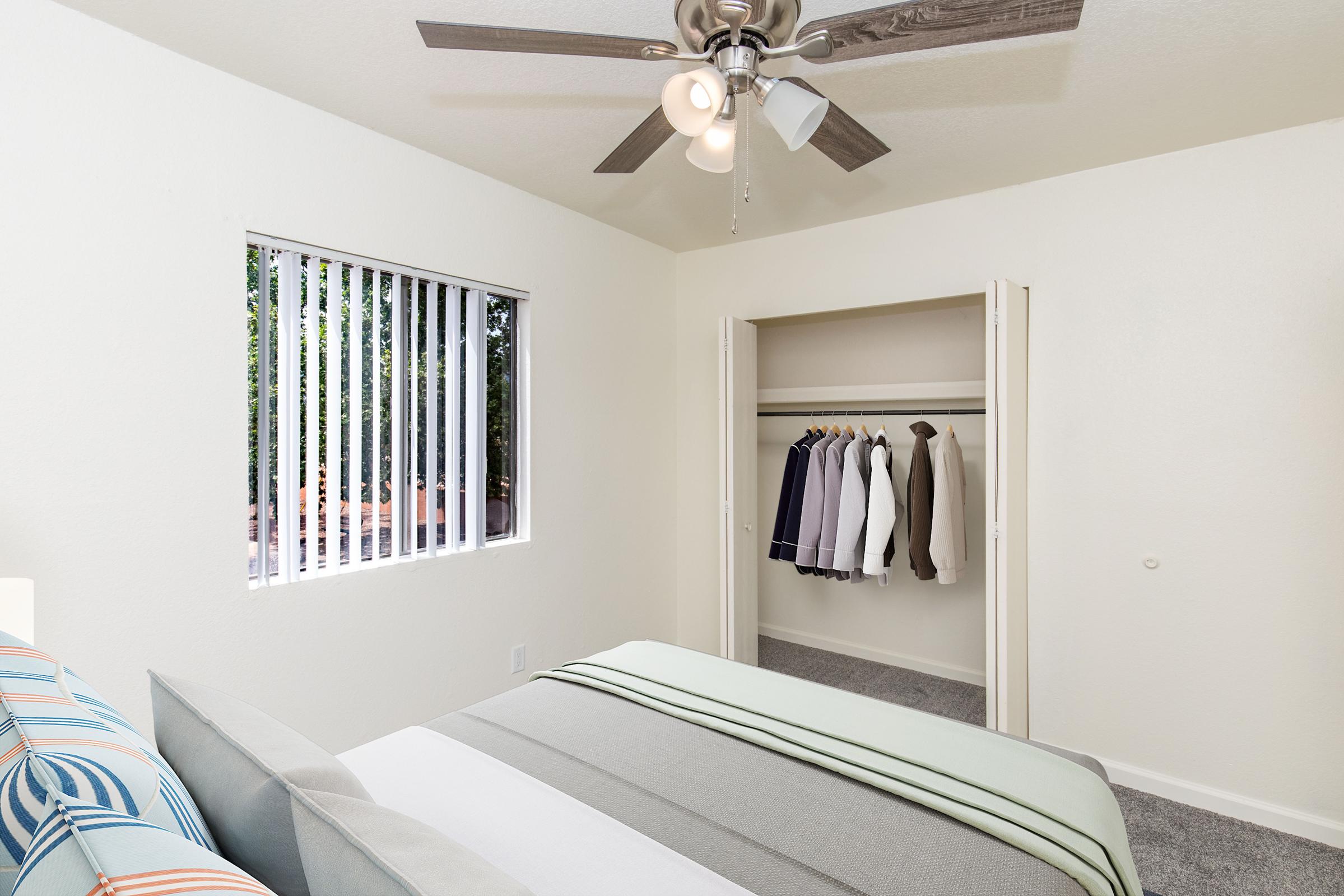 A well-lit bedroom featuring a ceiling fan, a cozy bed with striped bedding, and a closet with neatly arranged clothes. A window with vertical blinds allows natural light to enter, enhancing the inviting atmosphere of the space. The room has light-colored walls and a soft carpeted floor.