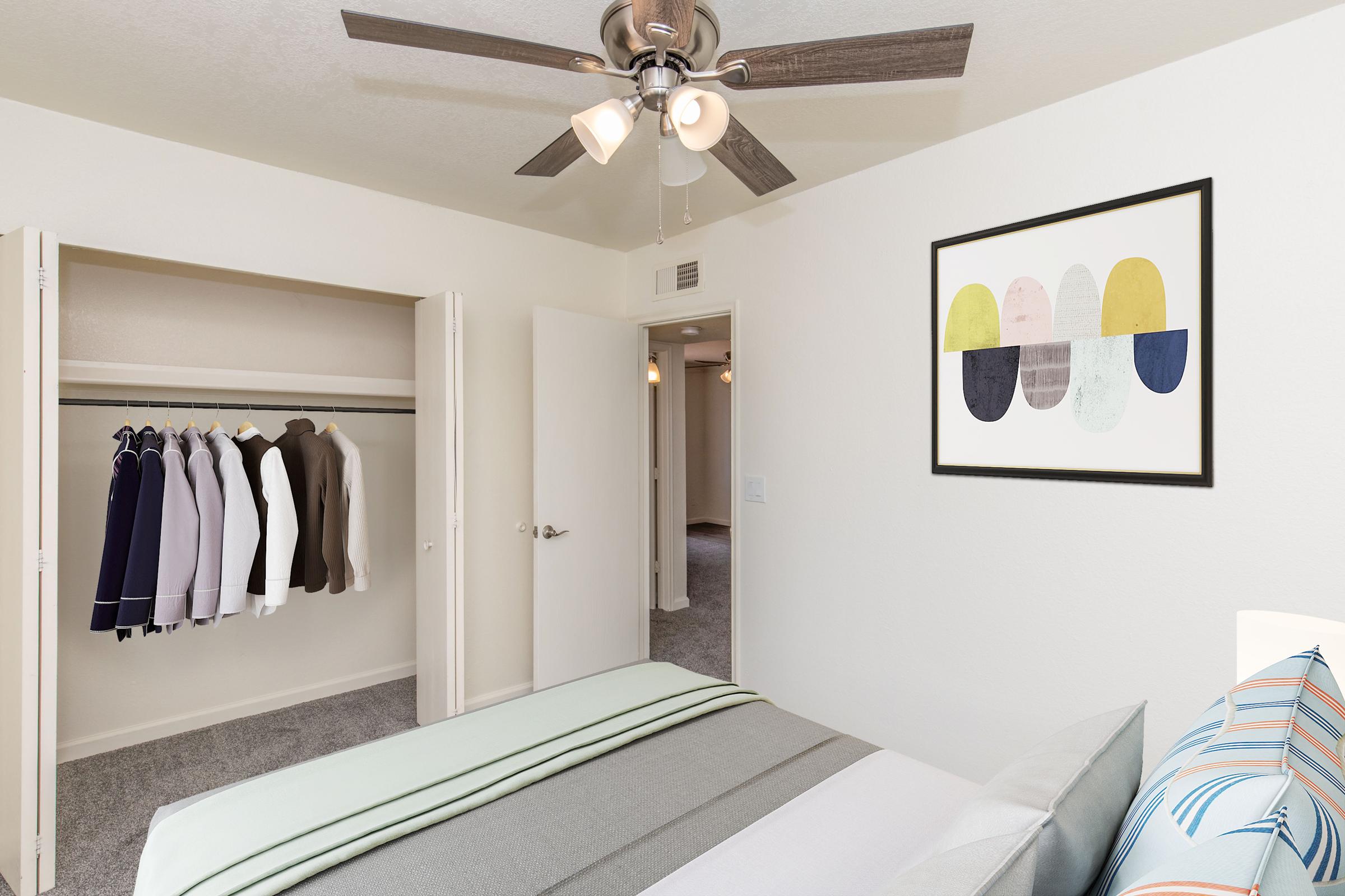 A well-lit bedroom featuring a bed with a light gray and white striped quilt. Adjacent to the bed is a closet with several neatly hung jackets. A door leads to another room, and a wall art piece with abstract shapes in muted colors hangs above the bed. The space has light-colored walls and a ceiling fan.