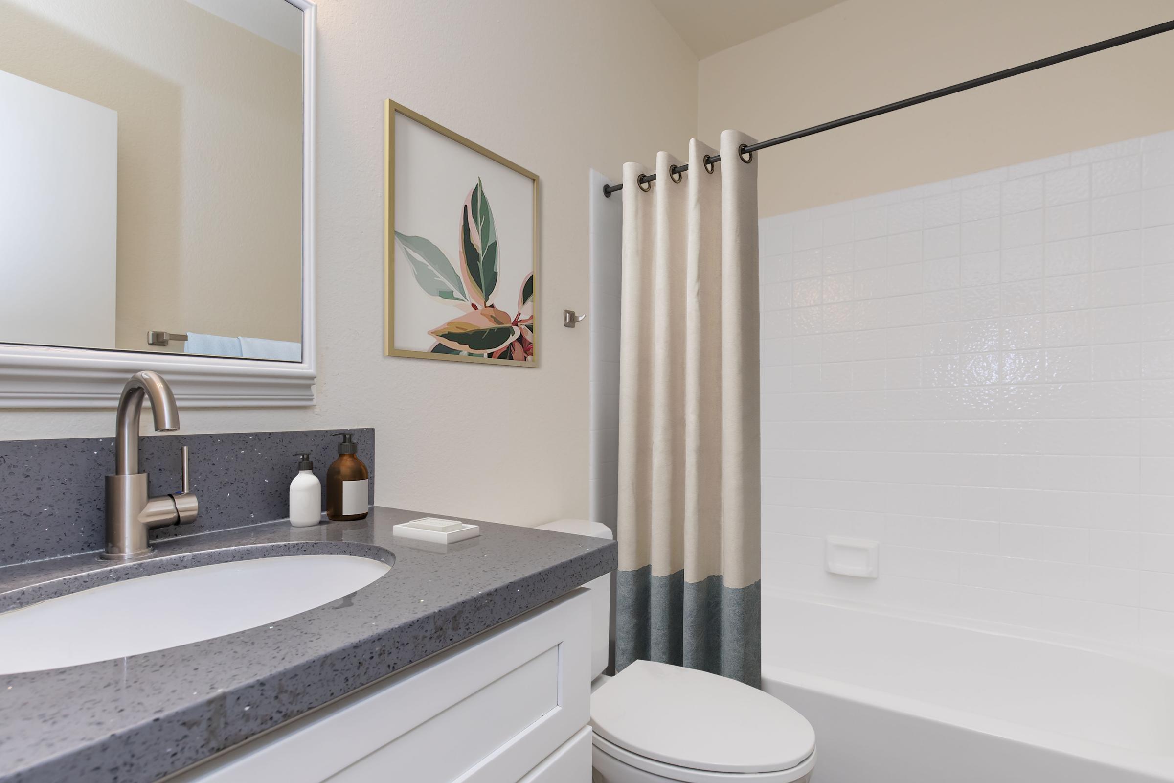 A modern bathroom featuring a clean white shower, gray countertop with a sink, and decorative items. A framed plant print decorates the wall, while a curtain in neutral tones provides privacy. The overall design is bright and contemporary, emphasizing simplicity and elegance.