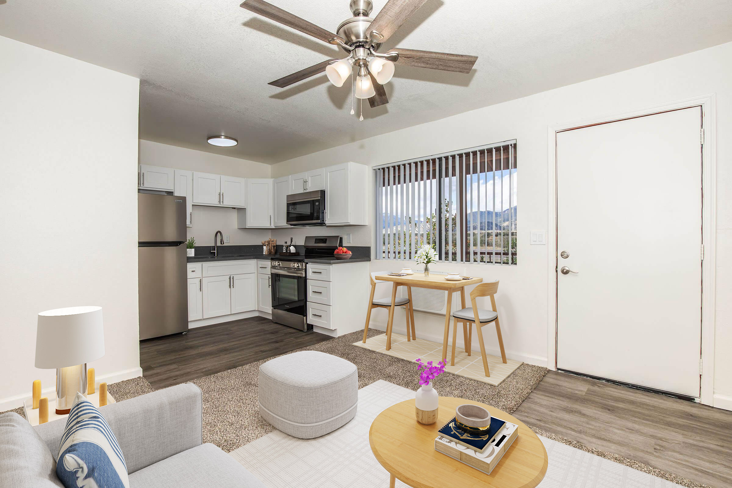 A modern kitchen and living area featuring white cabinets, stainless steel appliances, a dining table with two chairs, and a cozy sofa. Natural light streams through a window with blinds, revealing a mountain view. A ceiling fan hangs overhead, and a small coffee table displays a plant and decorative items.