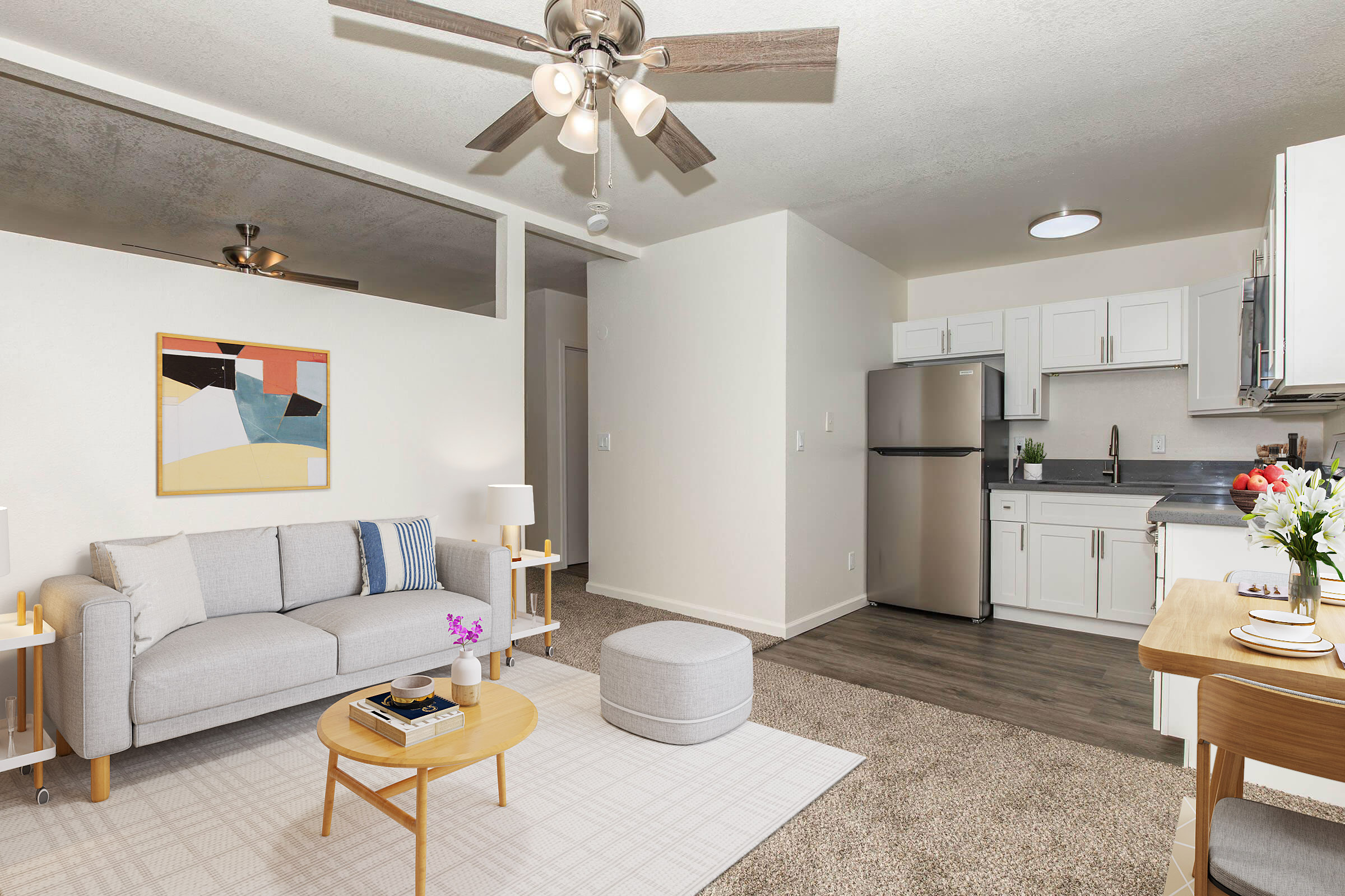 A modern living room and kitchen area featuring a light gray sofa, a round ottoman, and a wooden dining table set. Natural light illuminates the space, highlighting a ceiling fan and a minimalist decor. The kitchenette includes a silver refrigerator and white cabinetry, with a small vase of flowers on the table.