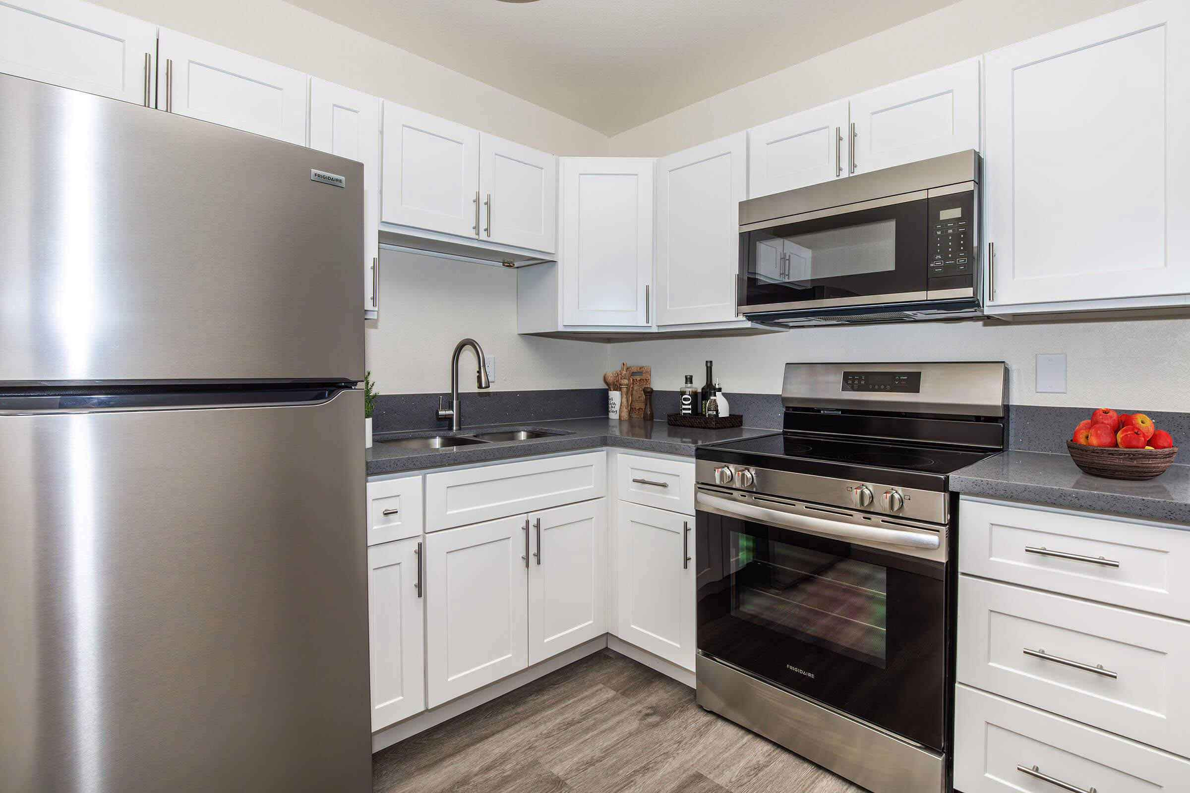 Modern kitchen with stainless steel appliances, including a refrigerator, microwave, and oven. White cabinetry provides storage and a sleek look. Gray countertops and a fruit bowl add a touch of color, while the overall layout is open and functional.