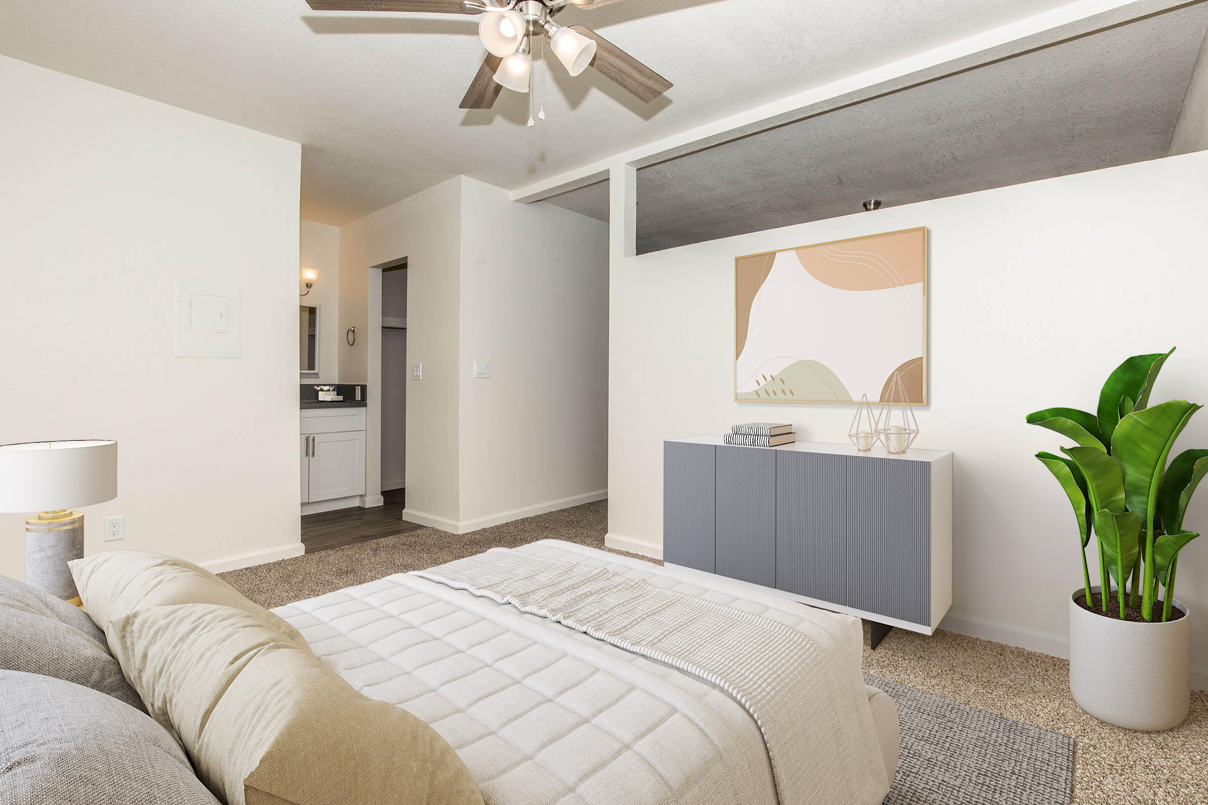 A stylish bedroom featuring a light-colored bed with decorative pillows, a modern dresser, a large plant, and a neutral wall art piece. A ceiling fan and soft carpeting create a cozy atmosphere. A doorway in the background leads to a bathroom area.