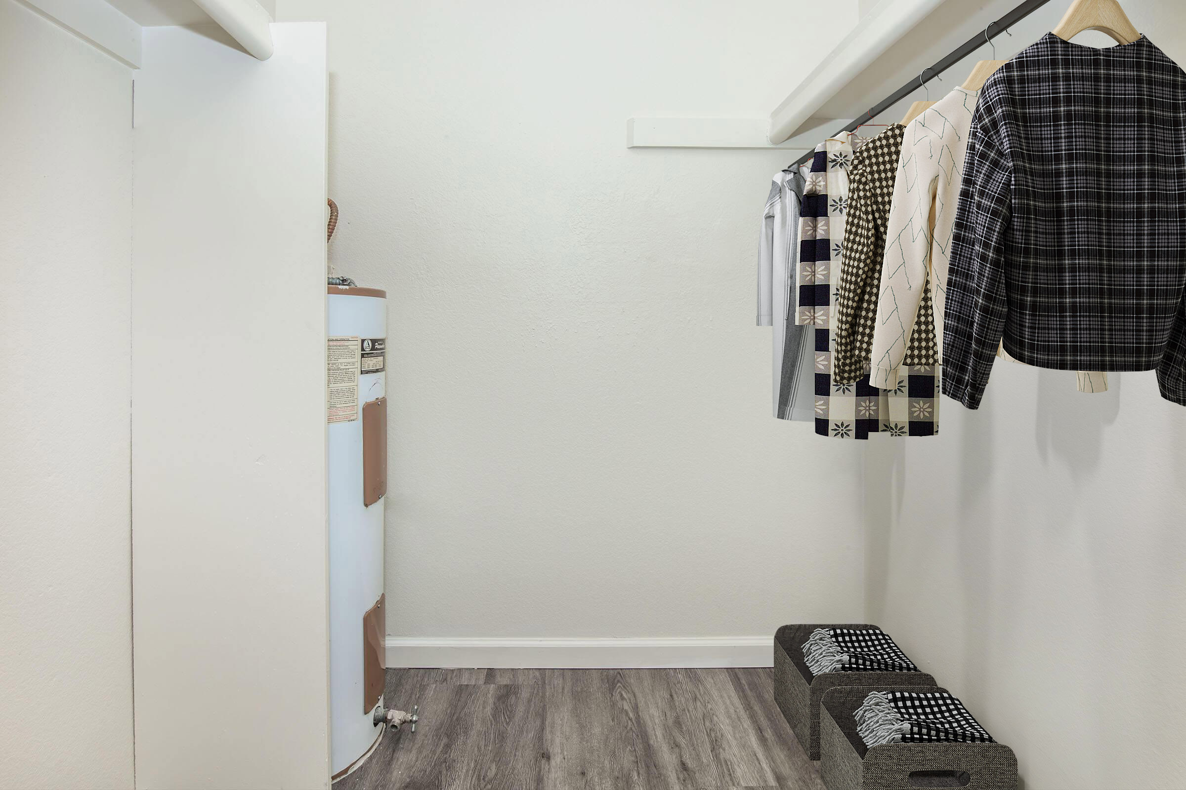 A small walk-in closet with white walls and a wooden floor. On the left, a white door leads outside. A white water heater is visible. To the right, several clothing items hang on a rail, including blouses and jackets. Two gray storage bins are placed on the floor, adding organization to the space.