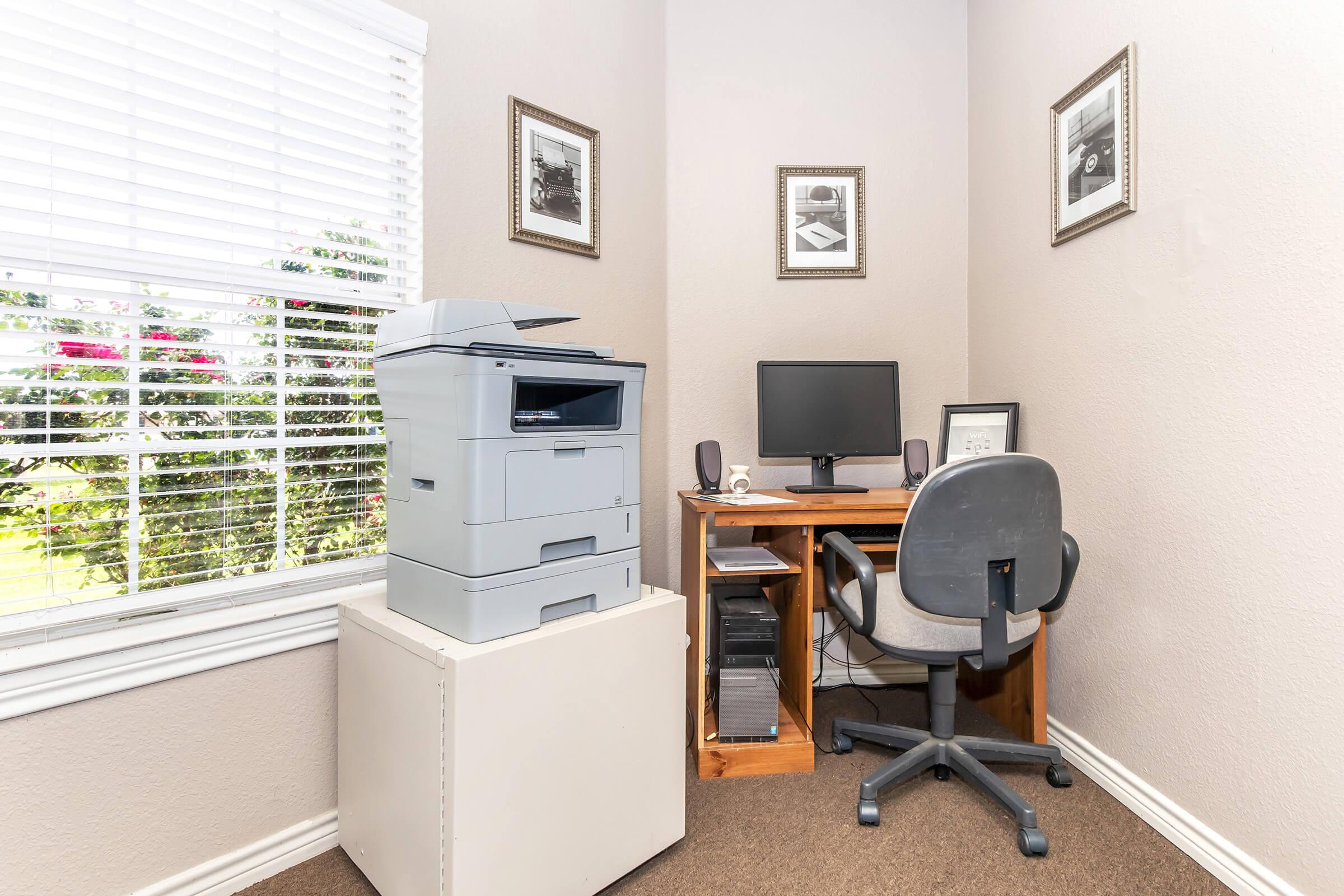 A small home office corner featuring a gray printer, a wooden desk with a computer monitor, and an office chair. The walls are painted in a light color, with framed photographs hanging. A window with white blinds allows natural light to enter, and leafy greenery is visible outside.