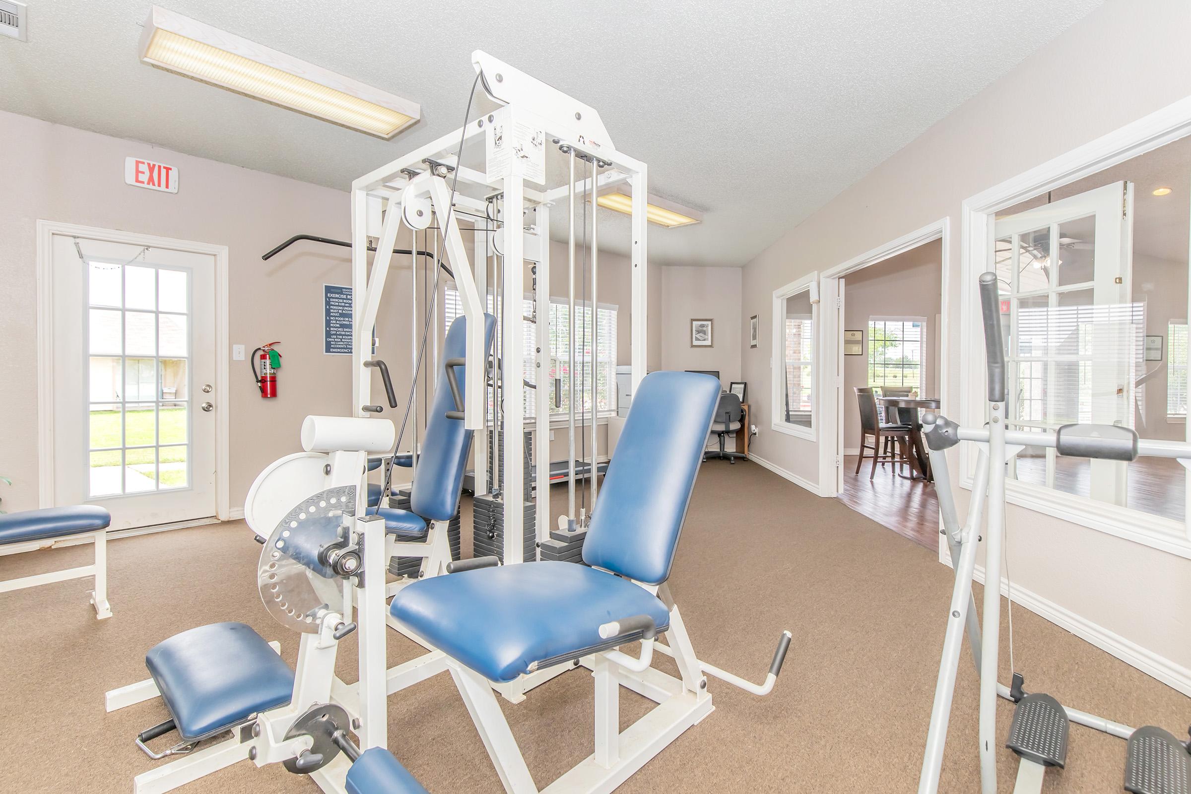 A well-lit gym room featuring a weight training machine with blue padded seats. The space includes another piece of equipment and has large windows allowing natural light. A door leads outside, and there are tables visible in an adjoining area. The walls are painted in neutral tones.