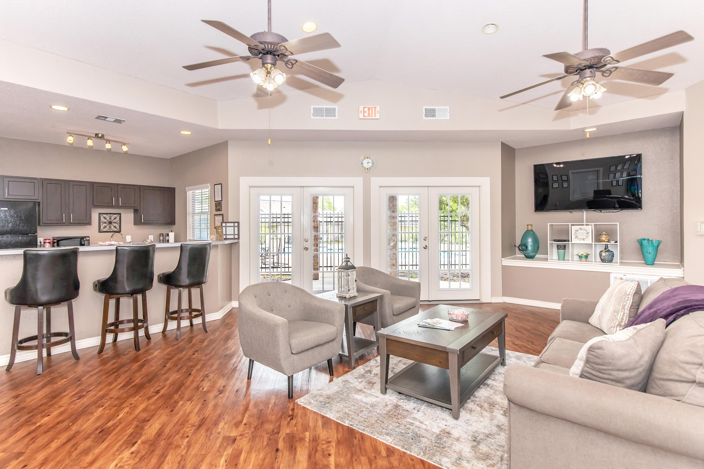 A modern living space featuring an open layout with hardwood floors, two ceiling fans, a comfortable sofa, chairs, and a coffee table. The kitchen area includes a breakfast bar with stools. Large windows open to a patio, and shelves display decorative items. Natural light brightens the room.