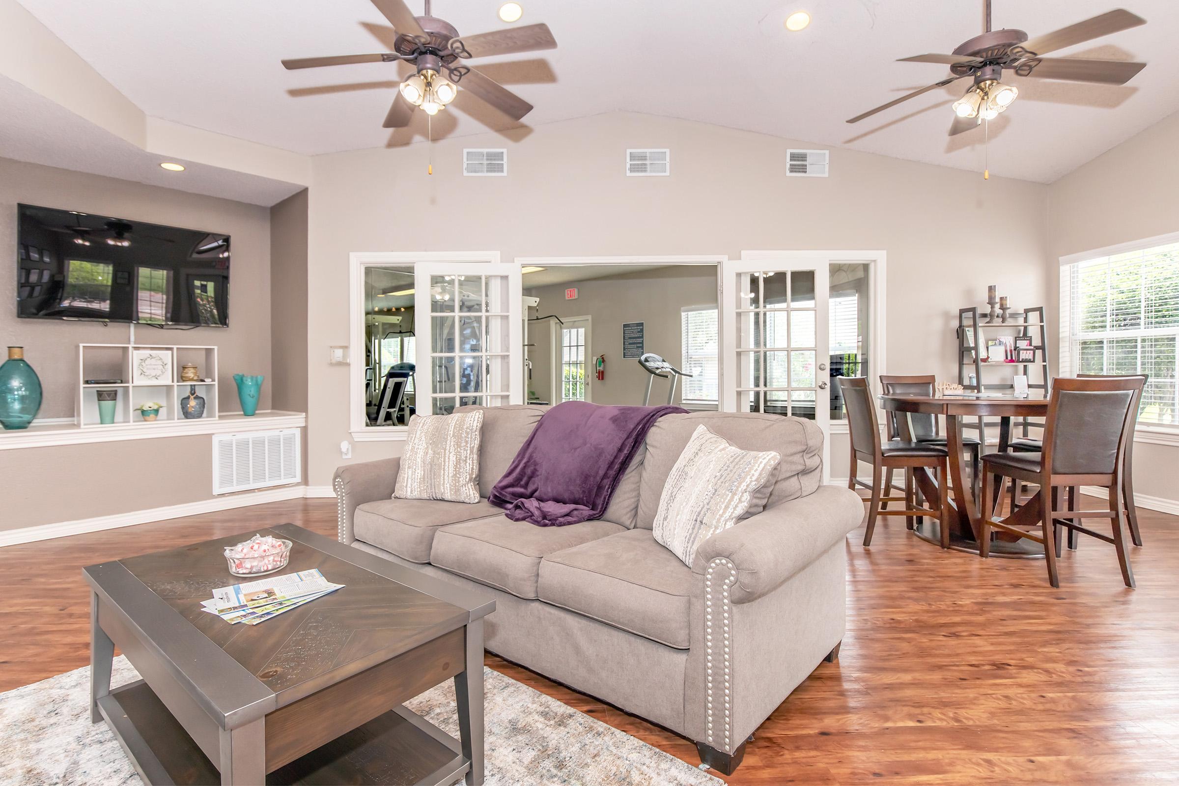 A spacious living room featuring a comfortable sofa with decorative pillows, a wooden coffee table, and a cozy rug. There are two ceiling fans above, large windows allowing natural light, and a dining area with a circular table and chairs in the background. Elegant decor pieces are displayed on shelves.