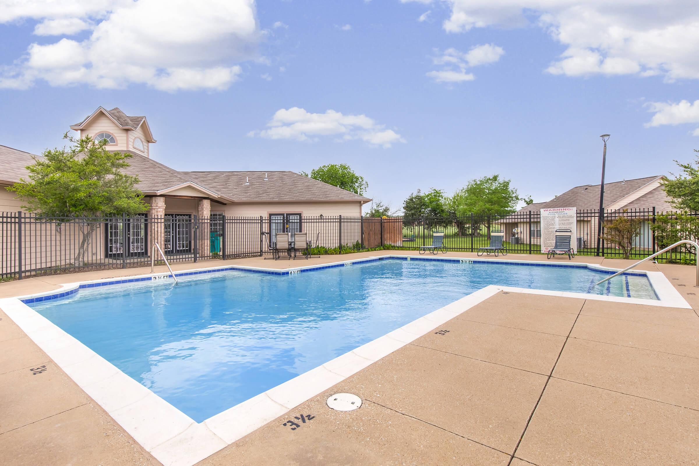 A clear, inviting swimming pool surrounded by a fenced area. There are lounge chairs positioned around the pool, with a light blue sky and a few trees in the background. The pool area is well-maintained, featuring a clean concrete deck and a white border around the water.