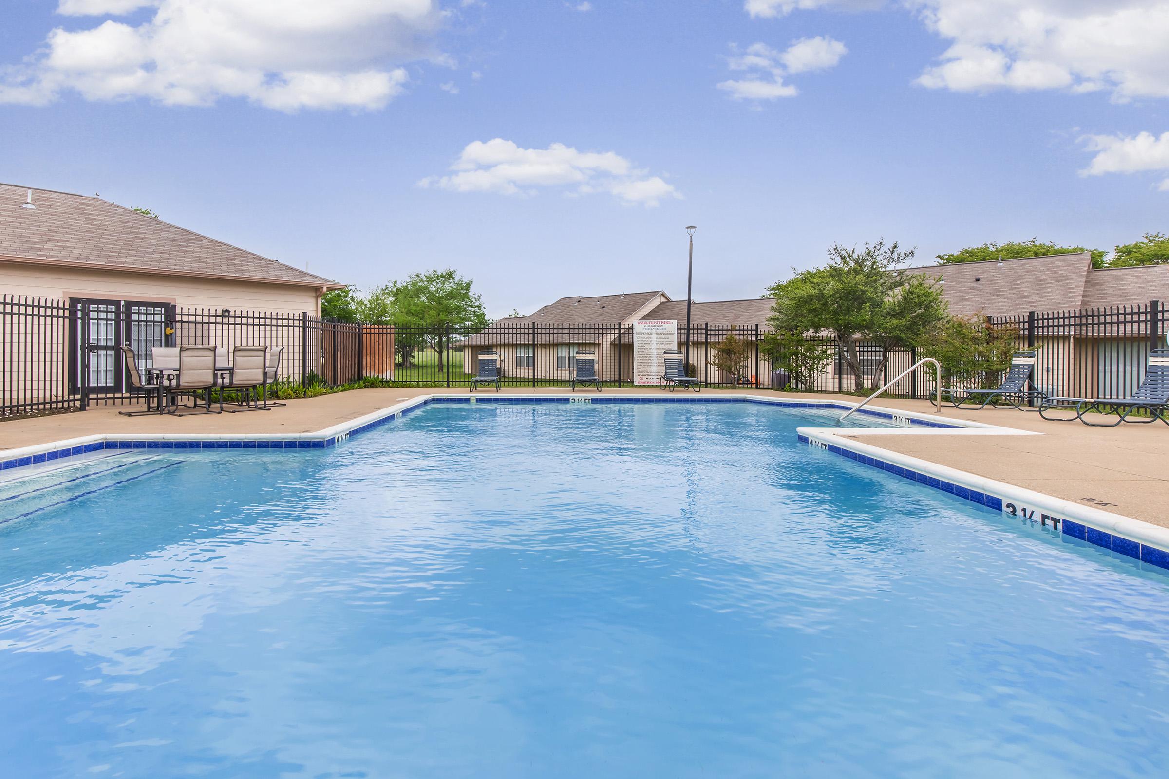 A serene swimming pool surrounded by a fence, with lounge chairs and tables placed nearby. The sky is clear with a few clouds, and residential buildings can be seen in the background. The water is crystal clear, reflecting the blue sky.