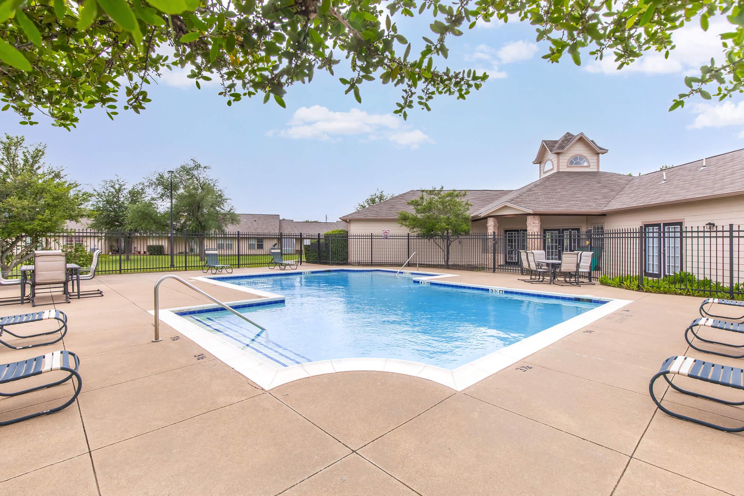 A clean and inviting outdoor swimming pool surrounded by lounge chairs, with a fence and landscaped greenery in the background. The sky is clear with a few clouds, and a building can be seen in the distance. The overall atmosphere is relaxed and perfect for recreation or leisure activities.