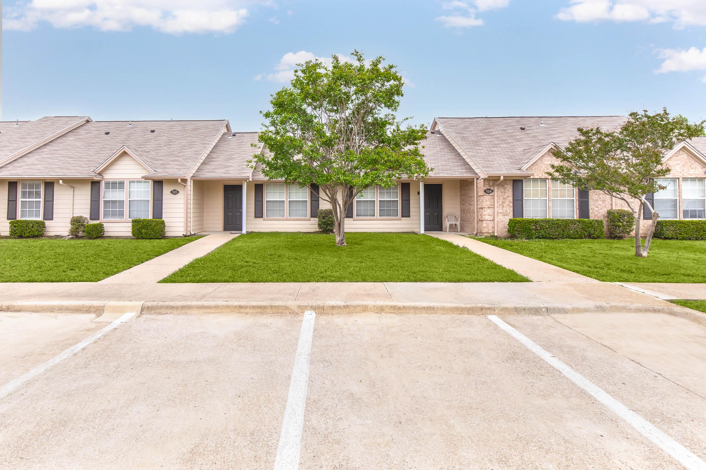 Row of single-story residential units with a grassy area in front. Each unit features a well-maintained lawn, walkways, and windows. A sizable tree is centrally located, providing shade. The scene is bright and inviting, showcasing a suburban neighborhood with parked cars in front.
