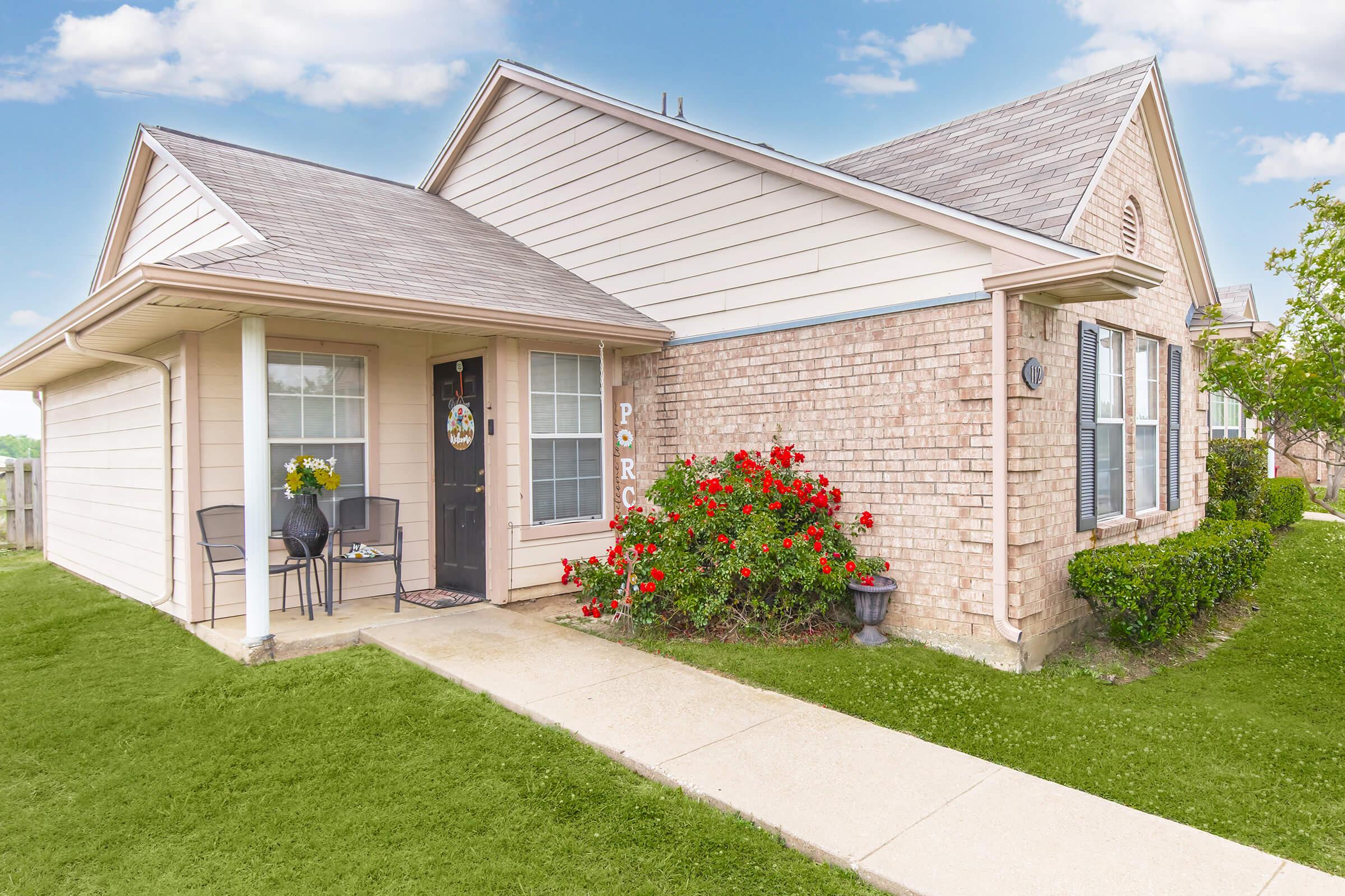 A cozy, single-story home with a light-colored exterior and brick accents. The front porch features a small table and chairs, surrounded by vibrant red flowers and well-maintained greenery. A clear blue sky and scattered clouds complete the welcoming outdoor scene.