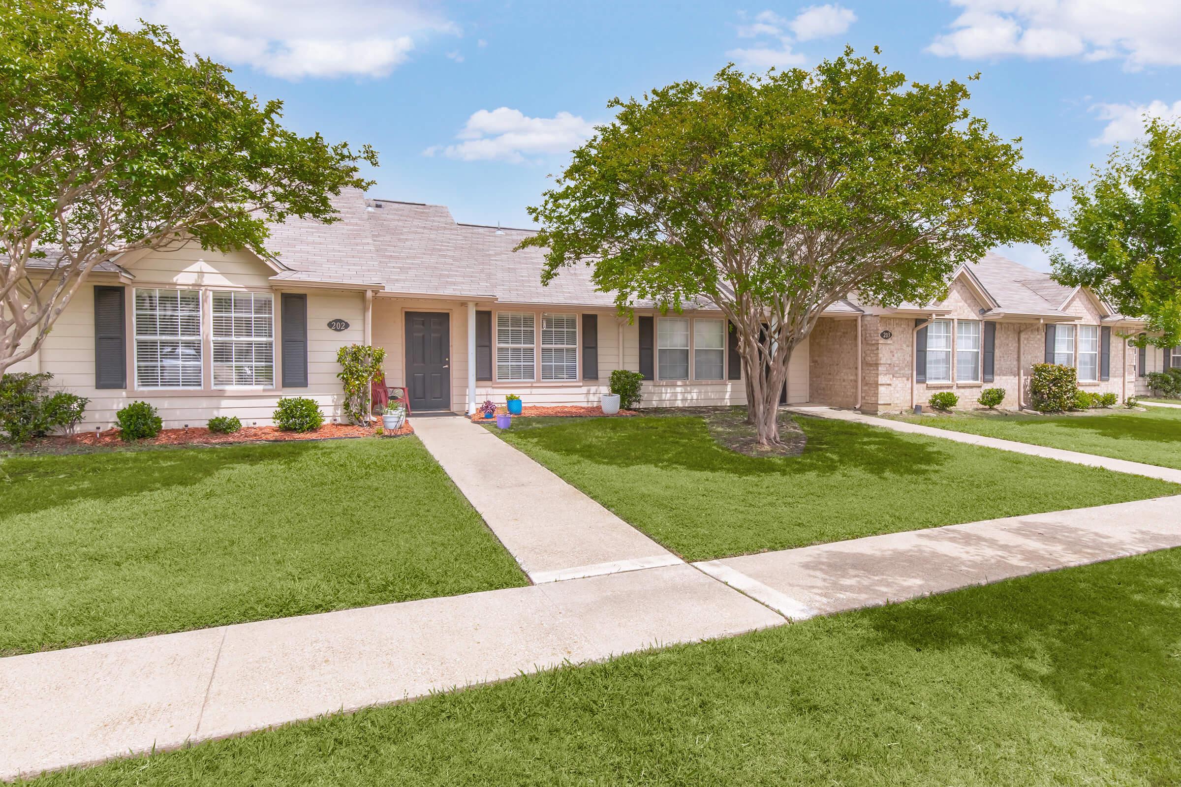 A well-maintained row of single-story houses set in a suburban neighborhood. Each house features a front porch, lawn, and shrubs, with a clear blue sky and fluffy clouds above. Sidewalks lead between the houses, providing a neat and inviting atmosphere.