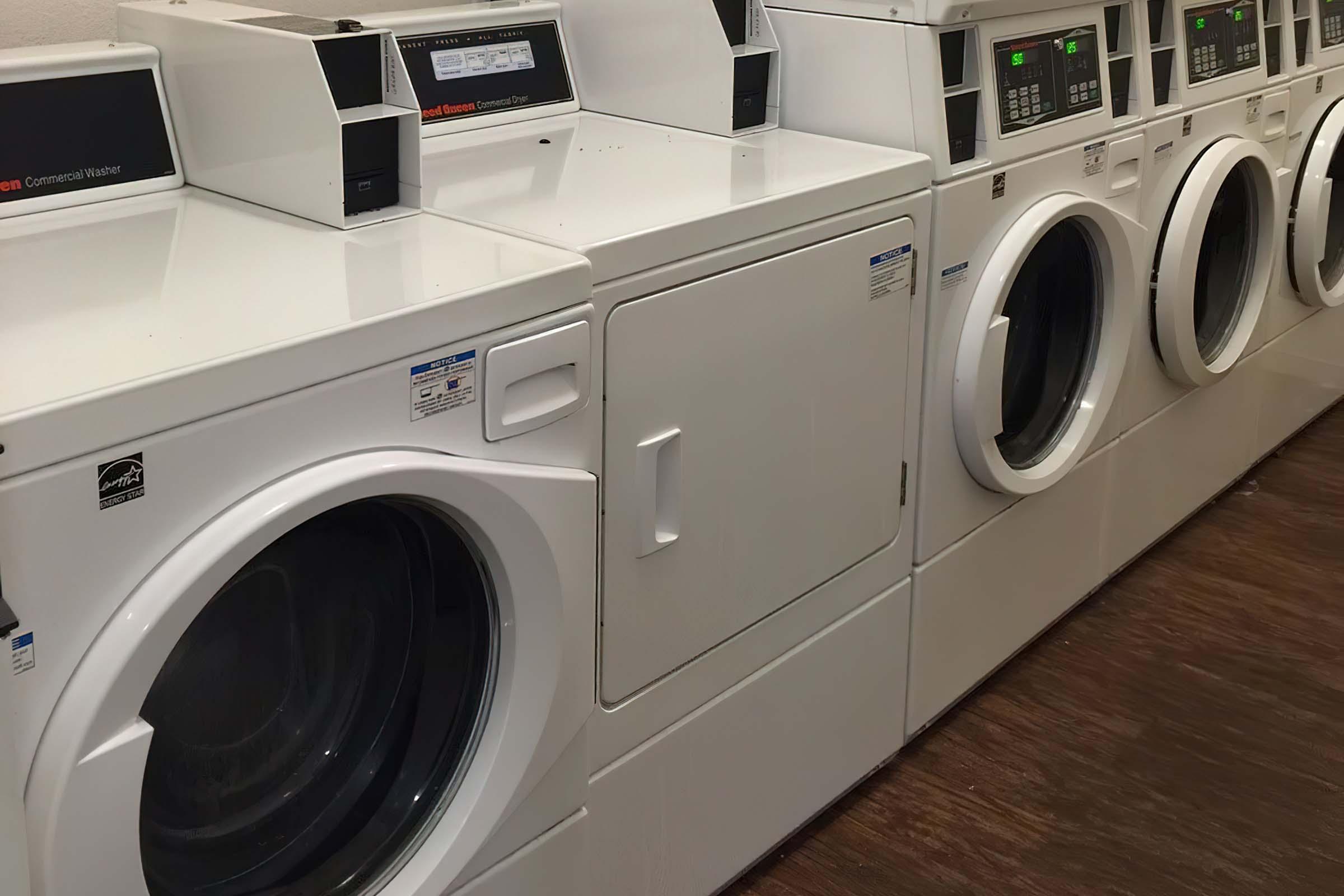 A row of white commercial washing machines and dryers in a laundromat, featuring digital control panels and large glass doors.