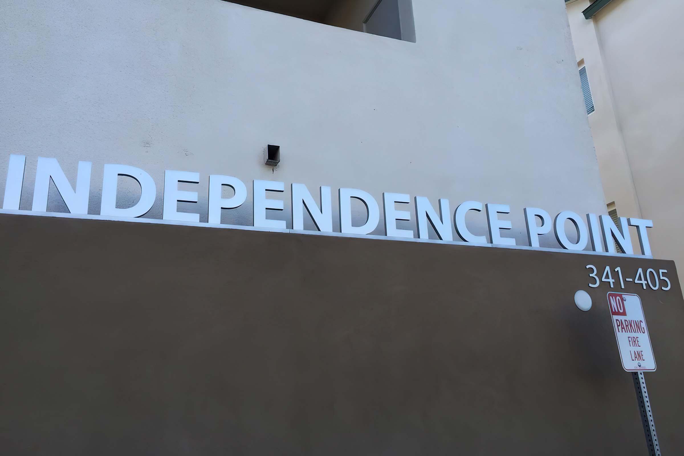 Sign displaying "INDEPENDENCE POINT" mounted on a wall, with a street sign below stating "NO PARKING FIRE LANE." The background features a beige wall.