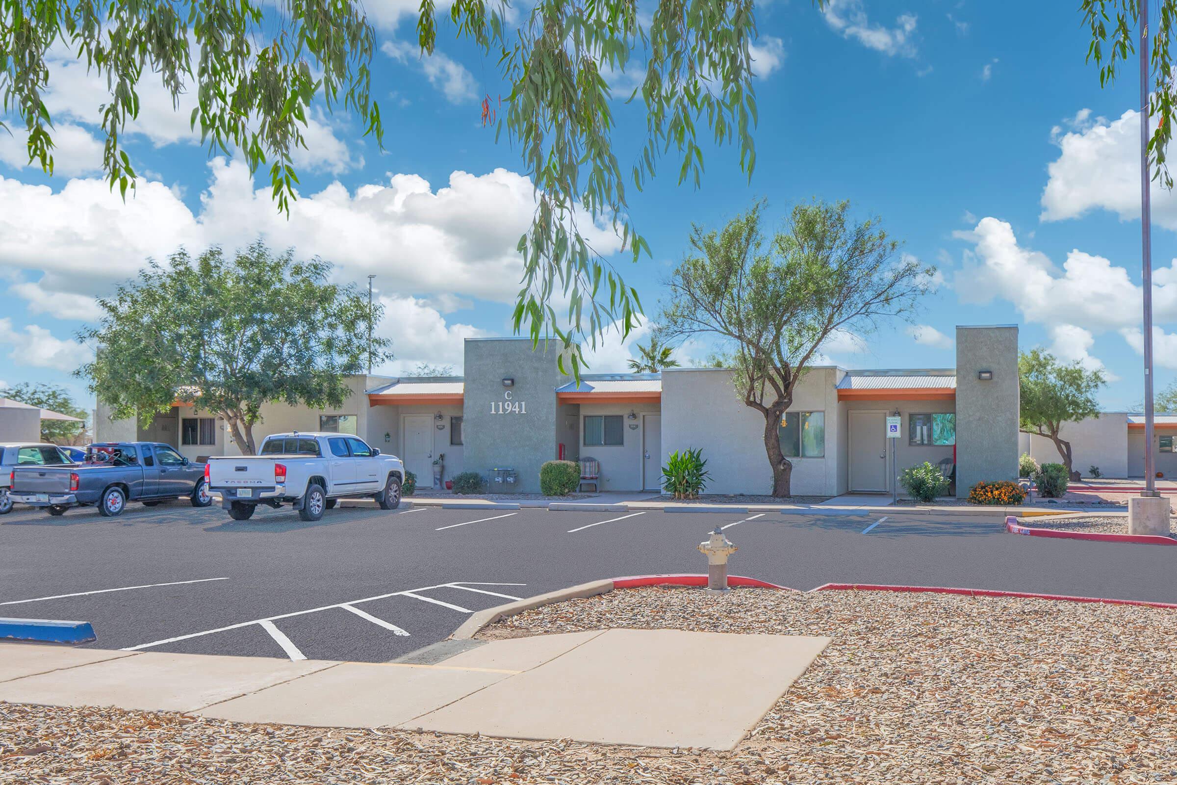 A view of a residential complex featuring modern buildings with flat roofs, surrounded by well-maintained landscaping. Vehicles are parked in the lot, and the sky is bright with fluffy clouds, creating a welcoming atmosphere.