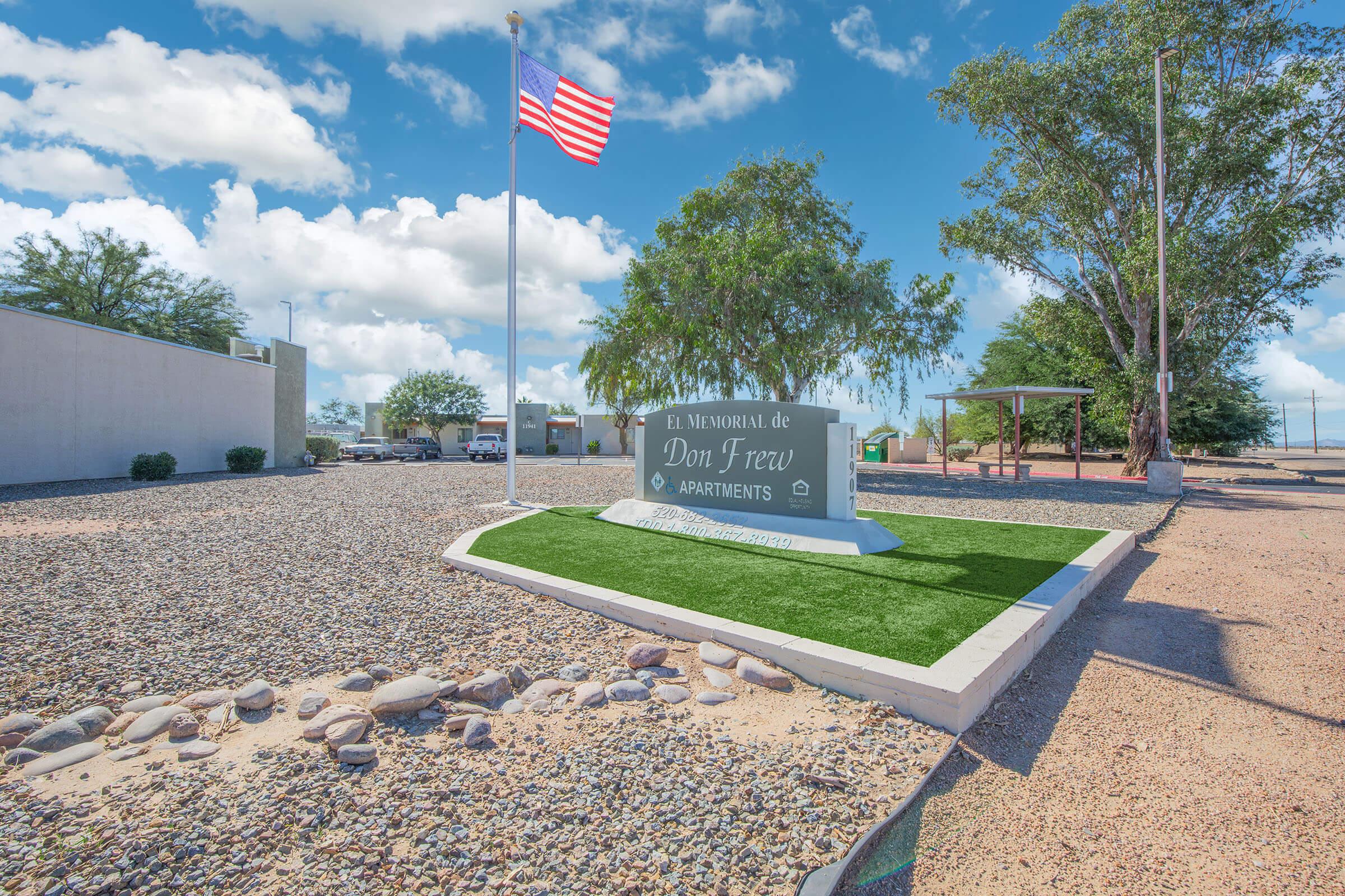 Sign for Don Frew Apartments located at a landscaped site with gravel and greenery, featuring an American flag on a pole in the background and partly cloudy blue skies.