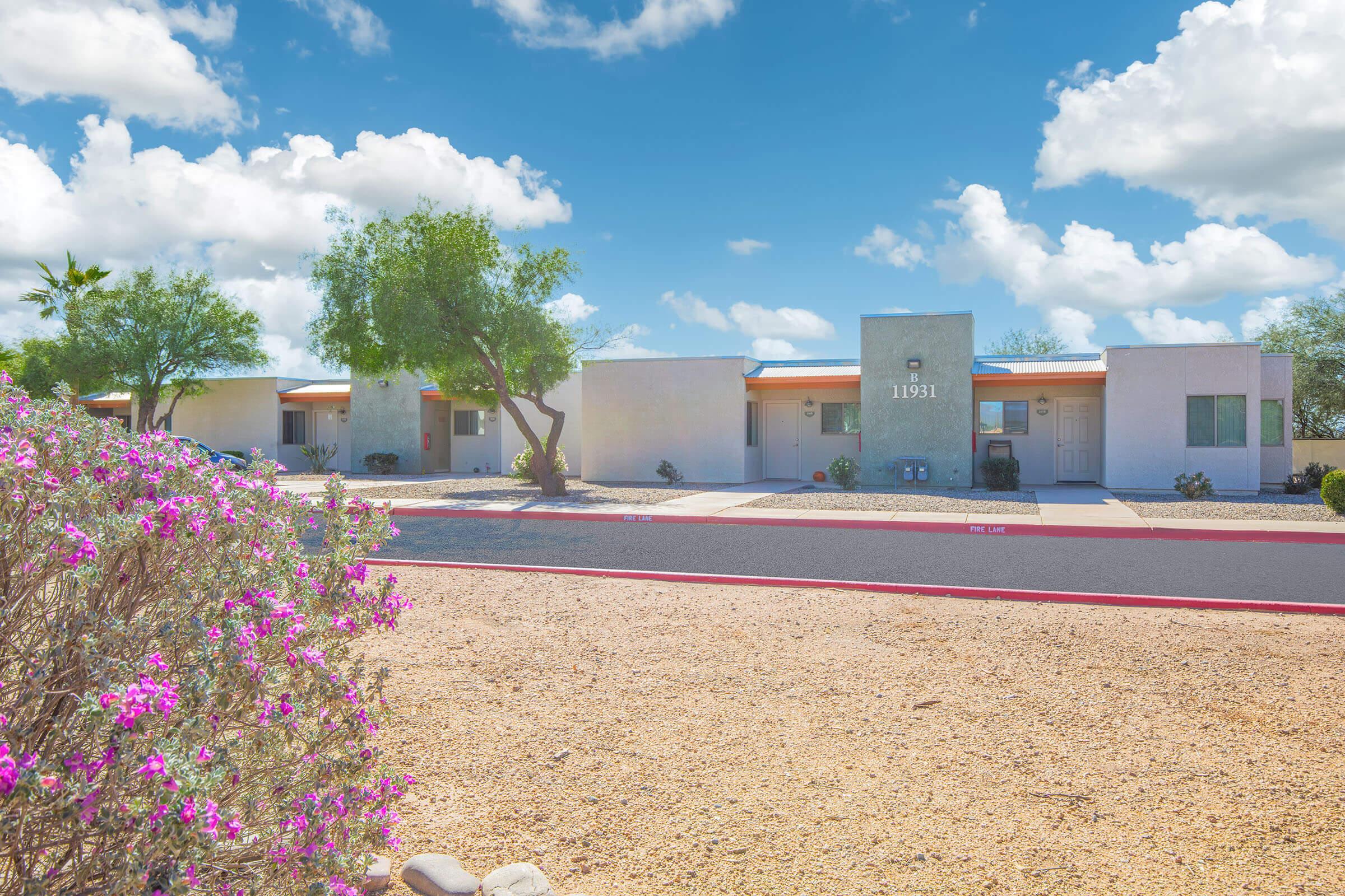 A sunny view of a residential complex featuring several single-story apartments. The buildings have light-colored walls and orange roofs, surrounded by desert landscaping with gravel and flowering plants in the foreground. A clear blue sky with fluffy white clouds complements the scene.