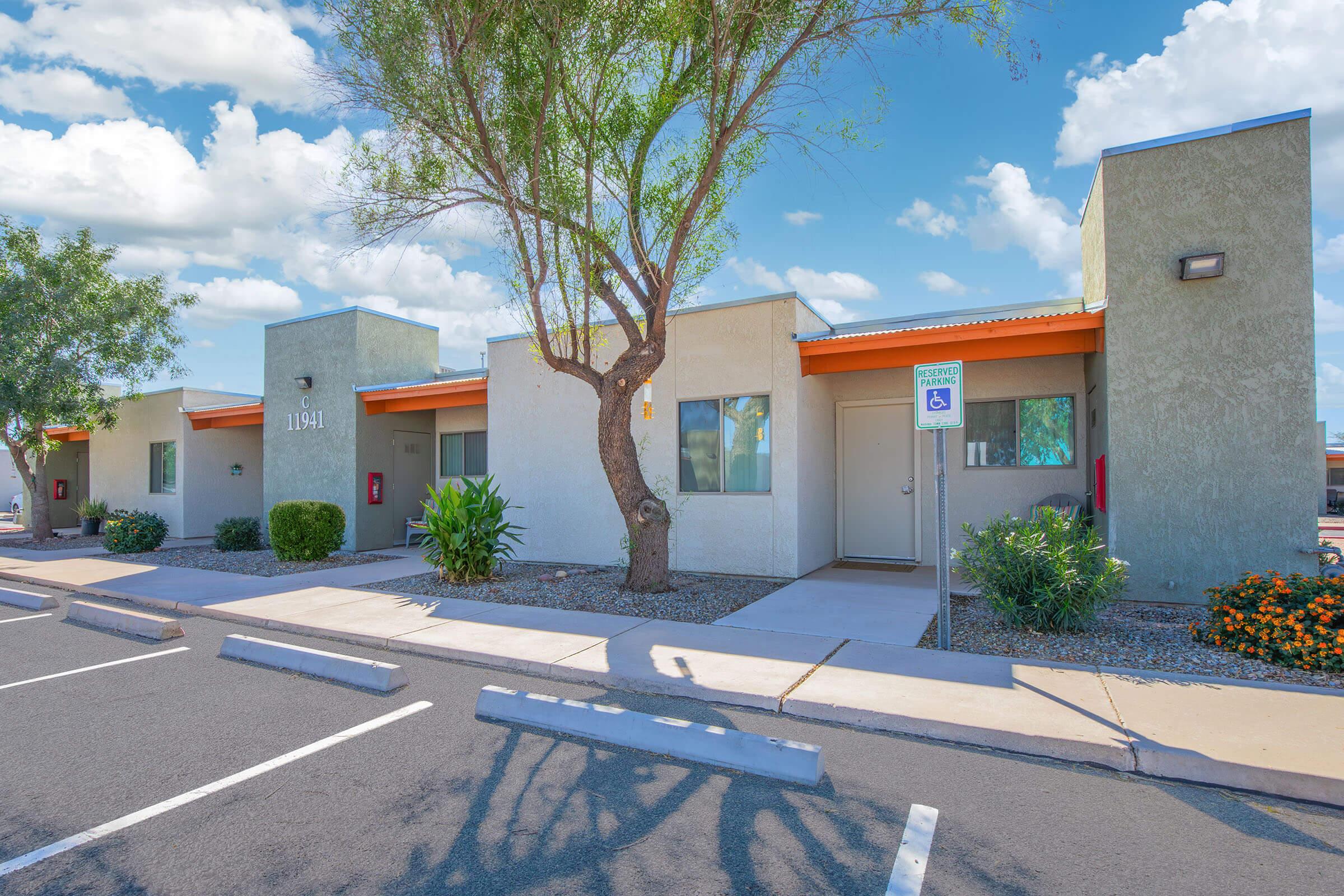 A modern, single-story building with a flat roof, featuring light-colored walls and orange accents. It has several windows, a tree in front, and well-maintained landscaping. There are parking spaces in front, and the sky is bright with fluffy clouds.