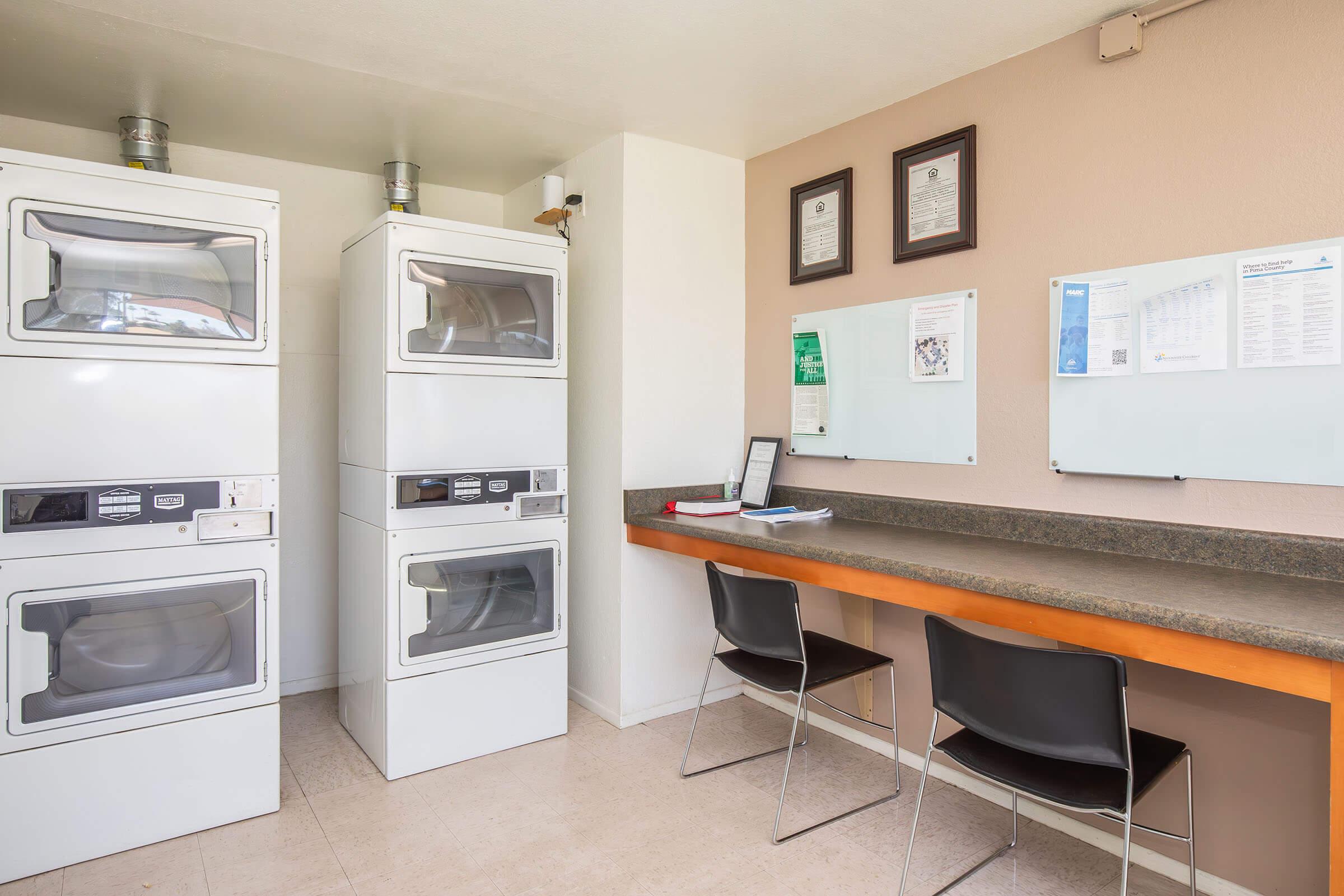A laundry room featuring two stacked washer-dryer units, a counter with two black chairs, and informational boards on the walls. The space is clean and well-lit, contributing to a functional and organized environment for laundry tasks.