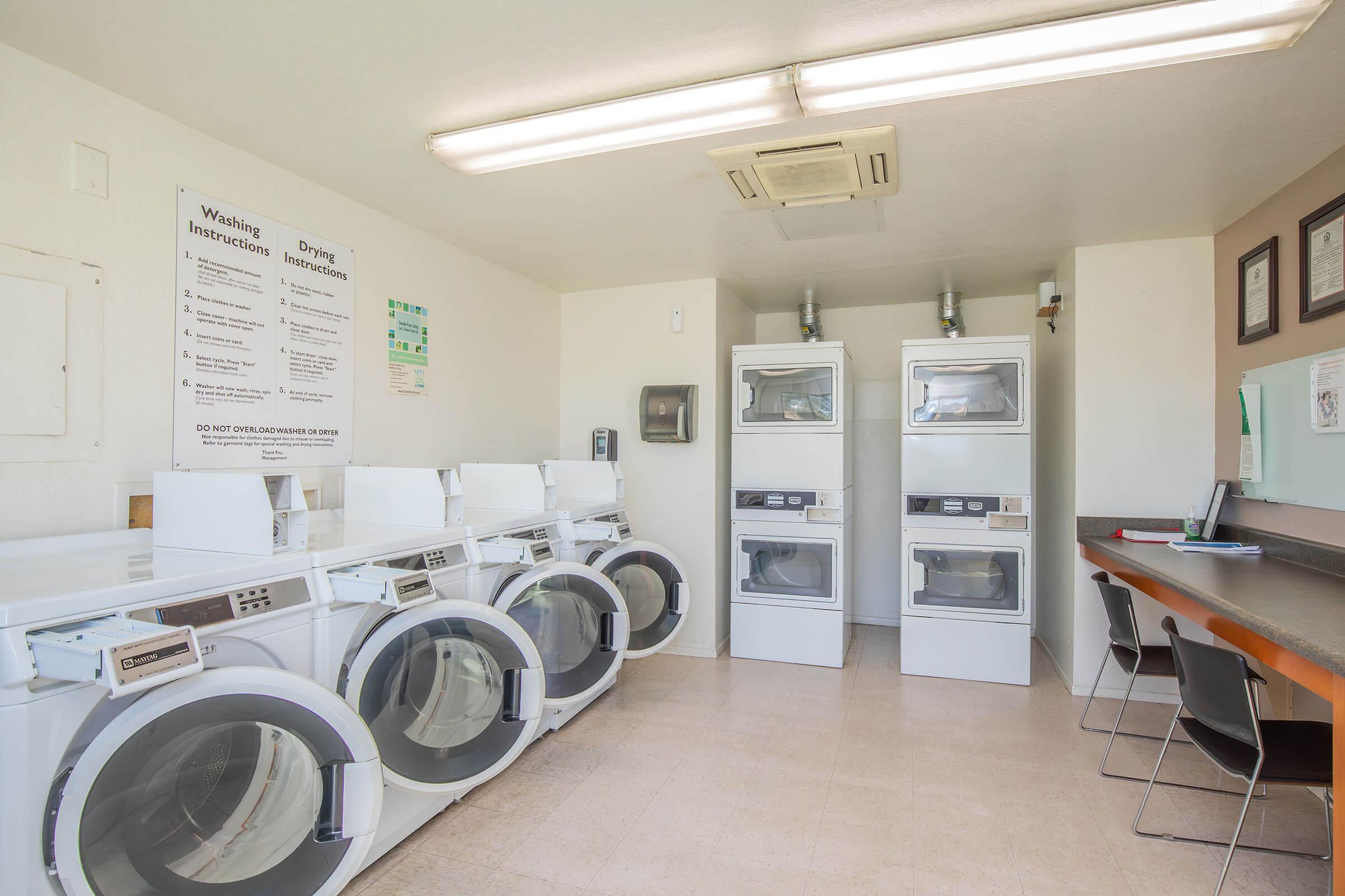 A clean laundry room featuring several washing machines and dryers. The machines are lined up against the wall, with instructional signs about washing and drying processes displayed. There's a counter with a couple of chairs and a wall-mounted information board. The space is well-lit and organized.