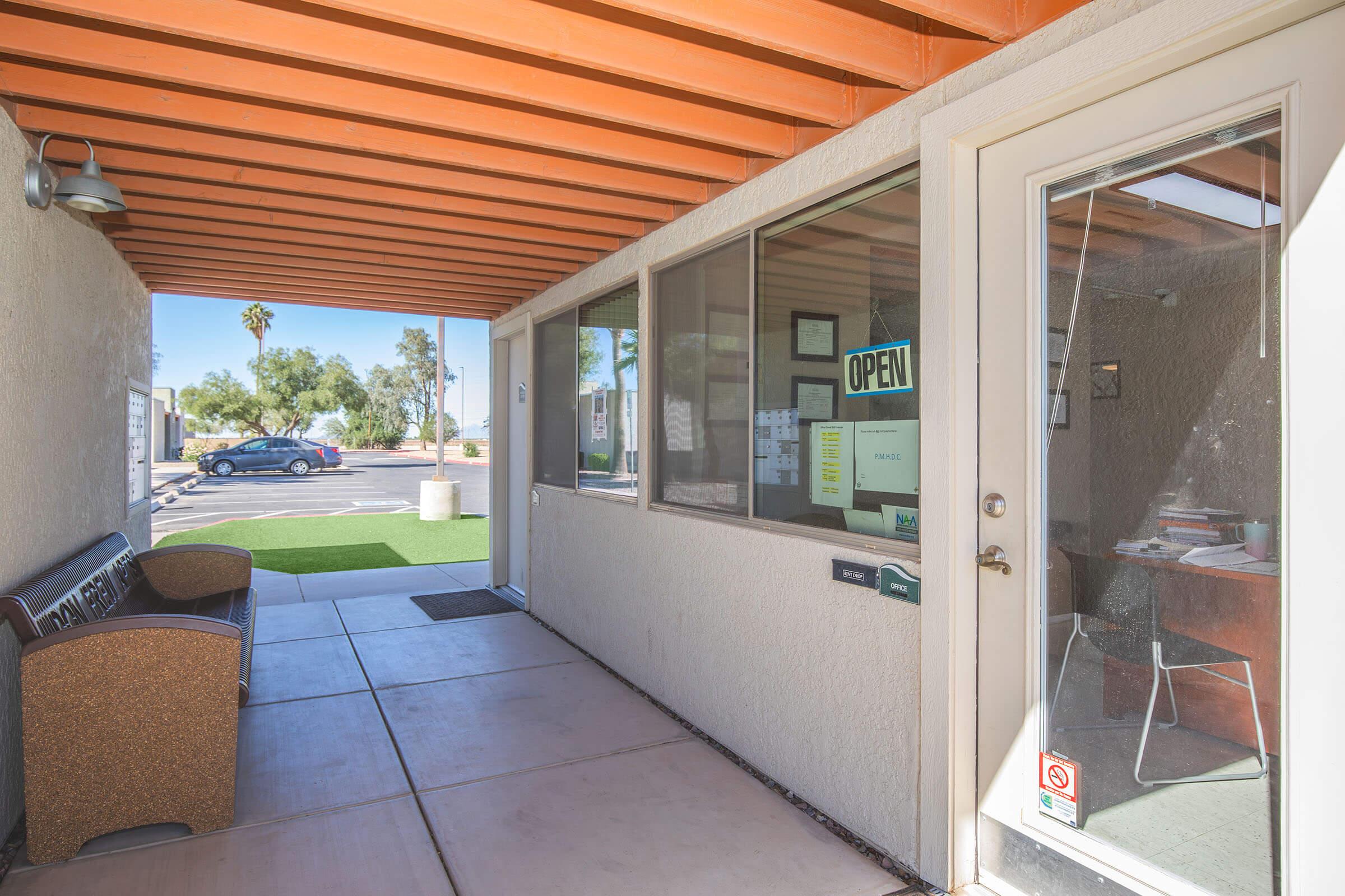A corridor with a glass door on one side showing an "OPEN" sign. There is a bench along the wall and a visible reception area with a desk and chairs. In the background, palm trees and vehicles are visible outside, suggesting a warm, inviting atmosphere.