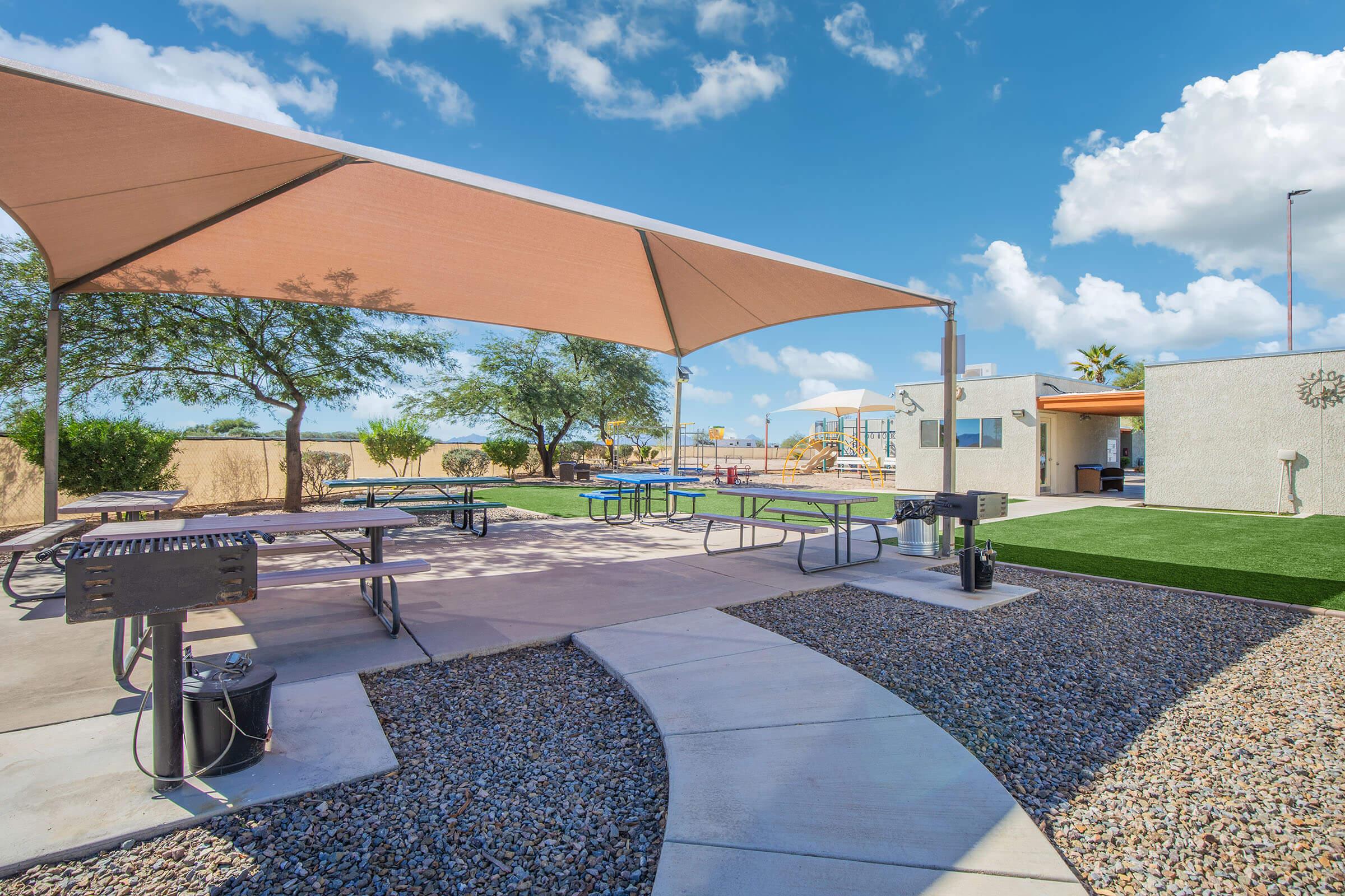 Outdoor recreational area featuring shaded picnic tables, gravel pathways, and green grass. In the background, there are trees and a playground. The sky is bright with fluffy clouds, creating a cheerful atmosphere. The space is designed for relaxation and social gatherings.