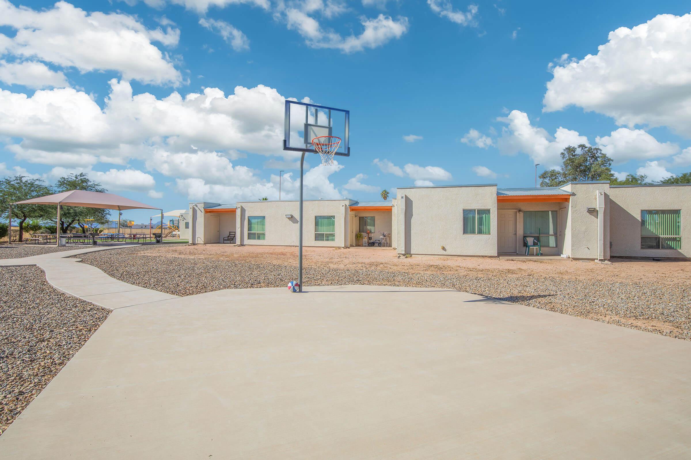 A secluded outdoor area featuring a basketball hoop on a concrete court, surrounded by gravel. In the background, there are several single-story buildings with orange accents and large windows under a bright blue sky with fluffy clouds. A shaded seating area is visible nearby.
