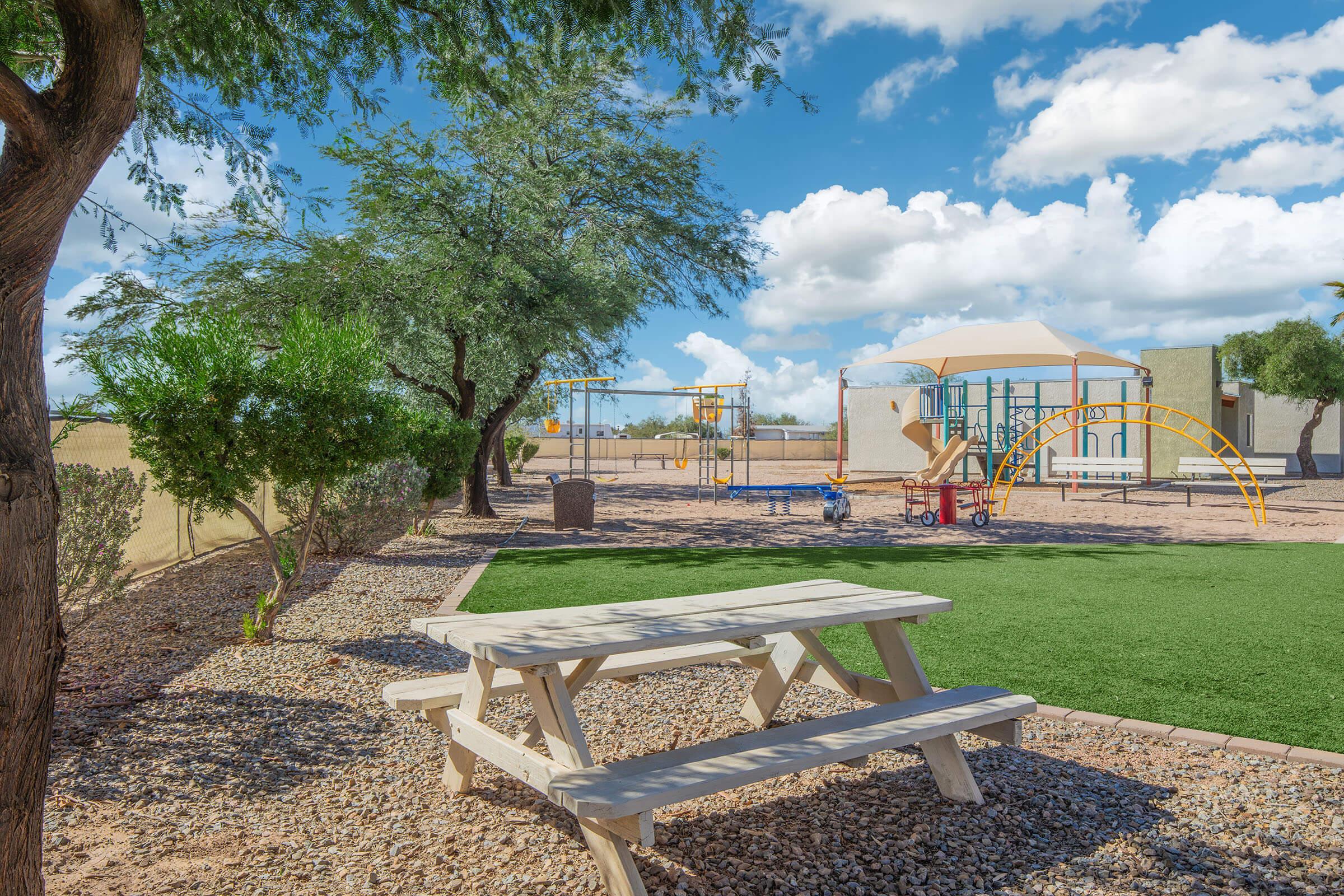 A children's playground featuring swings, slides, and climbing structures, surrounded by green grass and trees. A wooden picnic table is in the foreground, with a sandy area and blue sky filled with fluffy white clouds in the background.
