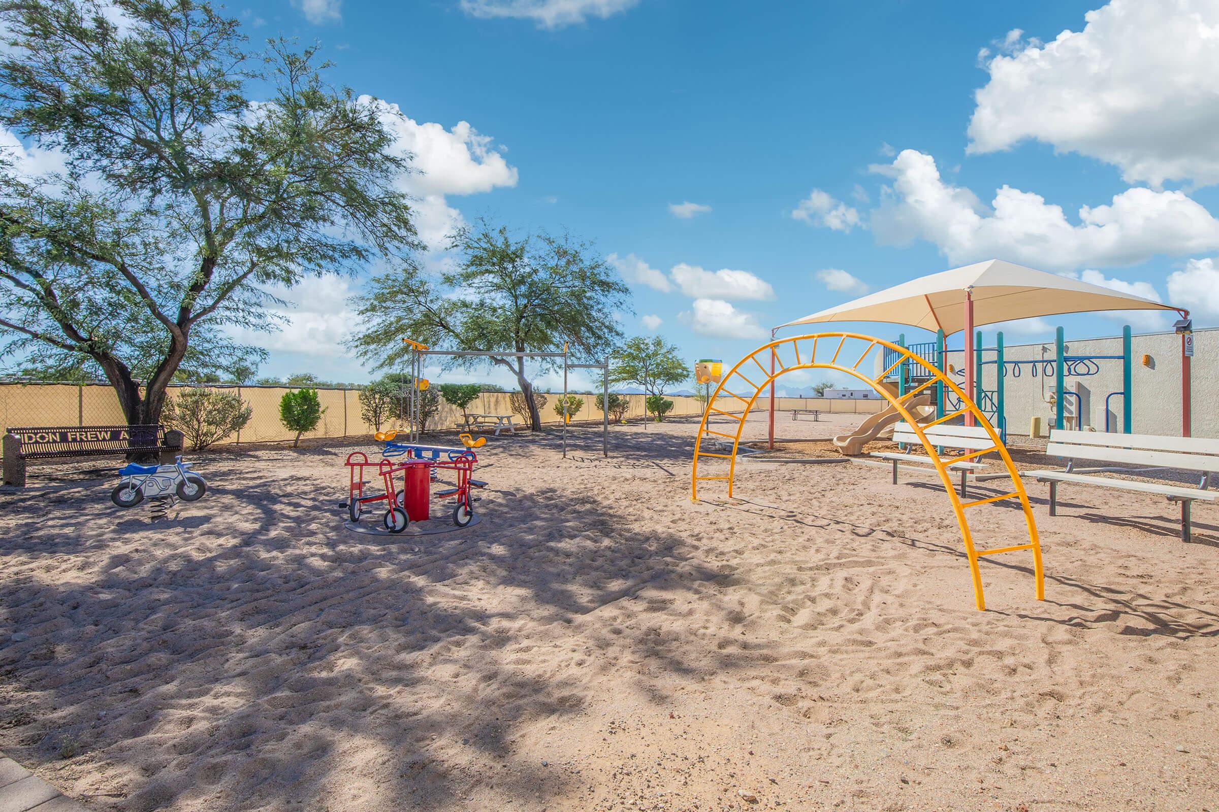 A playground featuring a sandy area with an arch-shaped climbing structure, a slide, and two tricycles parked nearby. Trees provide some shade, and a picnic table is visible in the background under a shade structure. The sky is bright with fluffy white clouds.