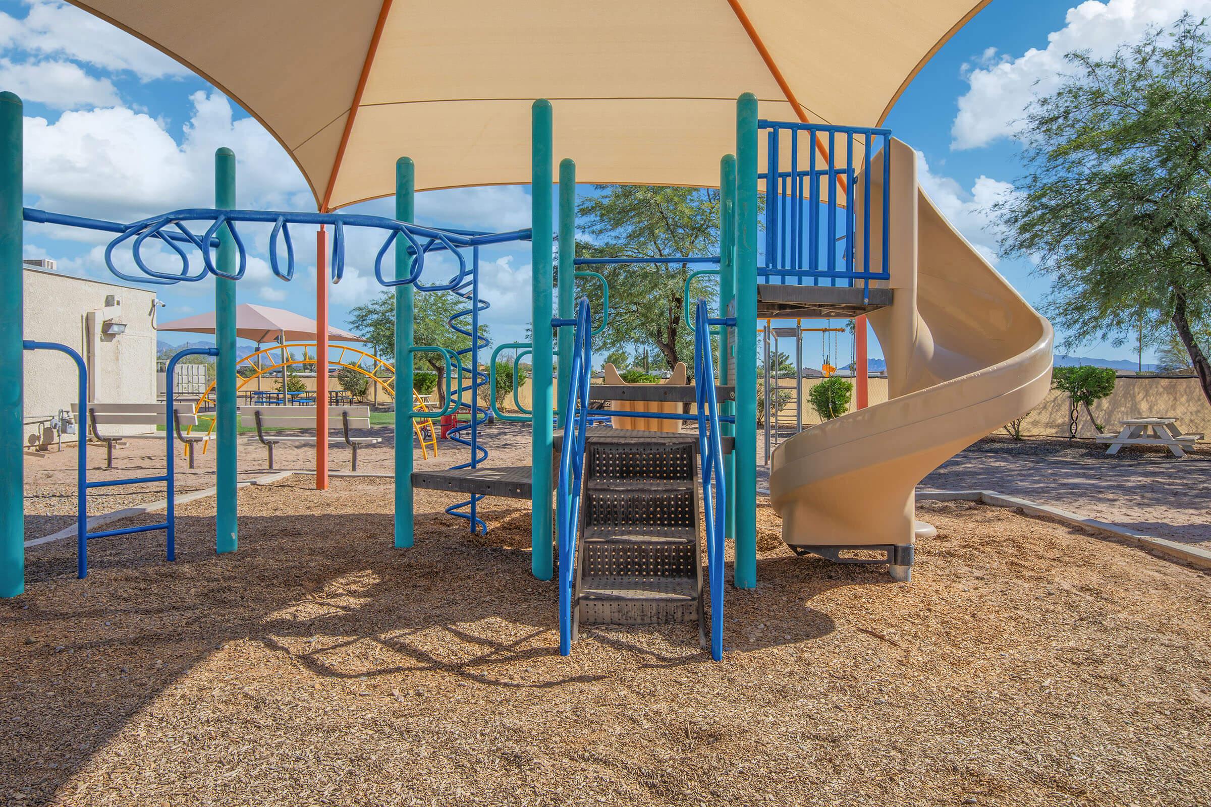 Playground equipment under a large shade canopy, featuring climbing apparatus, a spiral slide, and safety stairs, situated on a sandy surface with nearby trees and picnic tables in a sunny outdoor setting. Ideal for children's play and recreation.