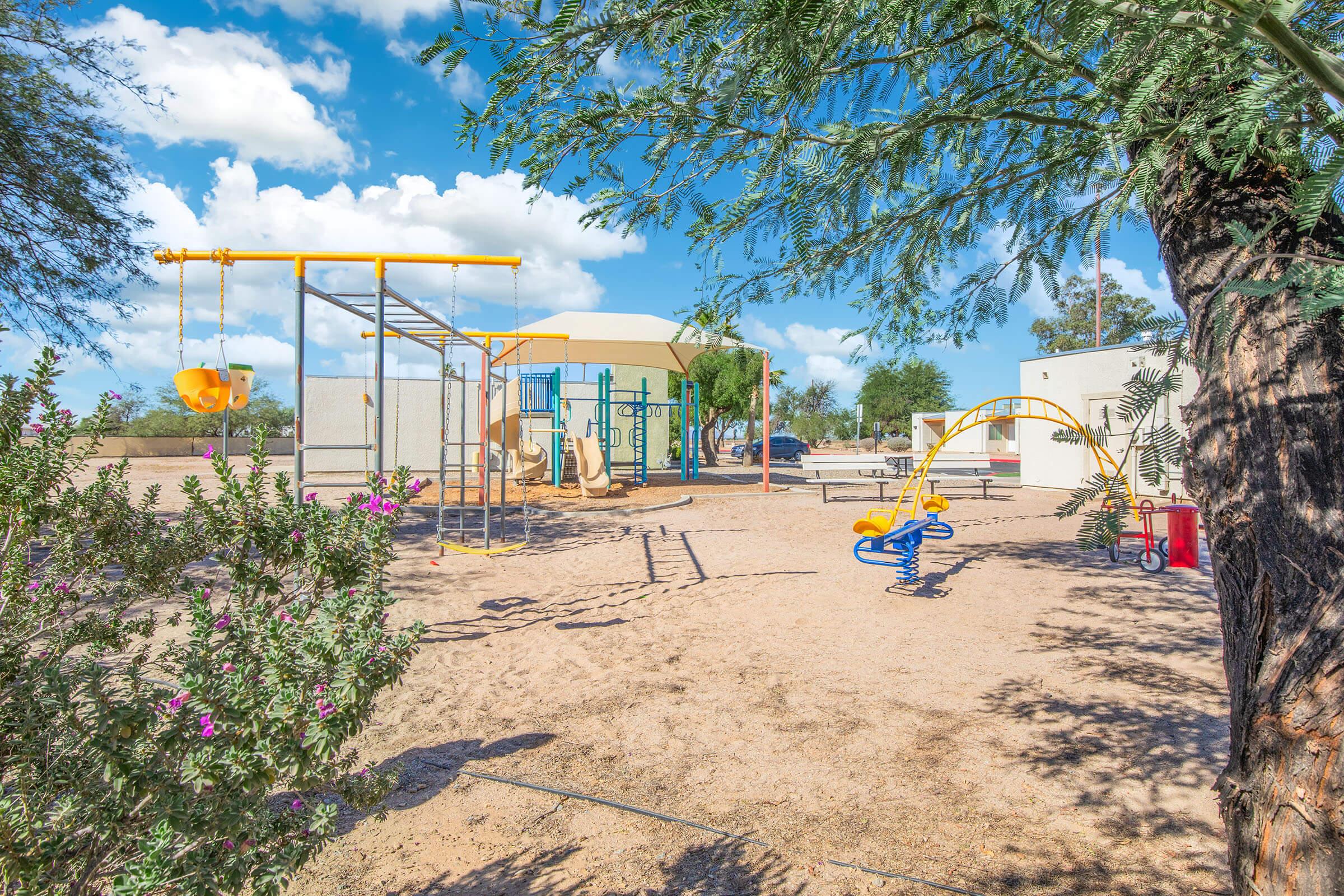 A playground with various equipment, including swings, slides, and climbing structures, surrounded by sand and greenery. The sky is clear with scattered clouds, and there are buildings in the background. The scene is bright and inviting, ideal for children to play and explore.