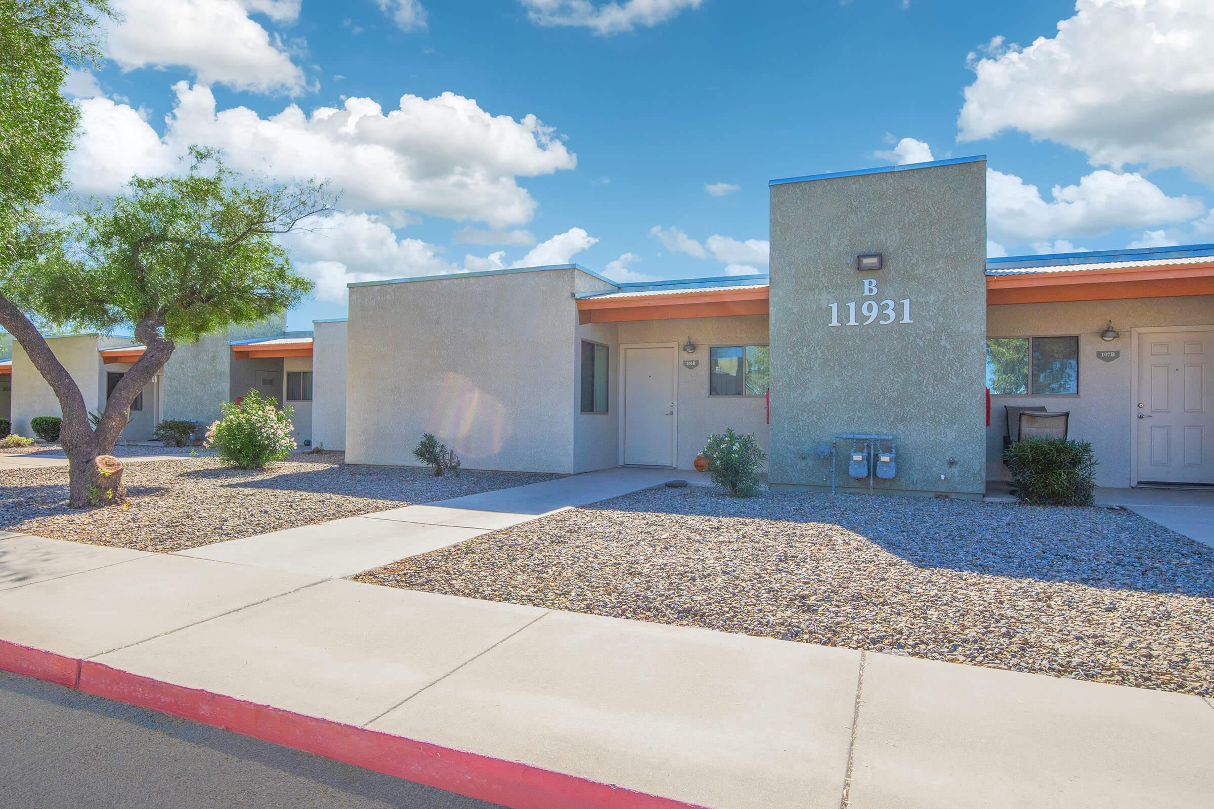 A single-story apartment building with multiple units, featuring a concrete walkway and landscaped areas with shrubs. The building has a light gray exterior with a blue sky and fluffy clouds overhead. The unit labeled 'B 11931' is visible, alongside a tree providing shade.