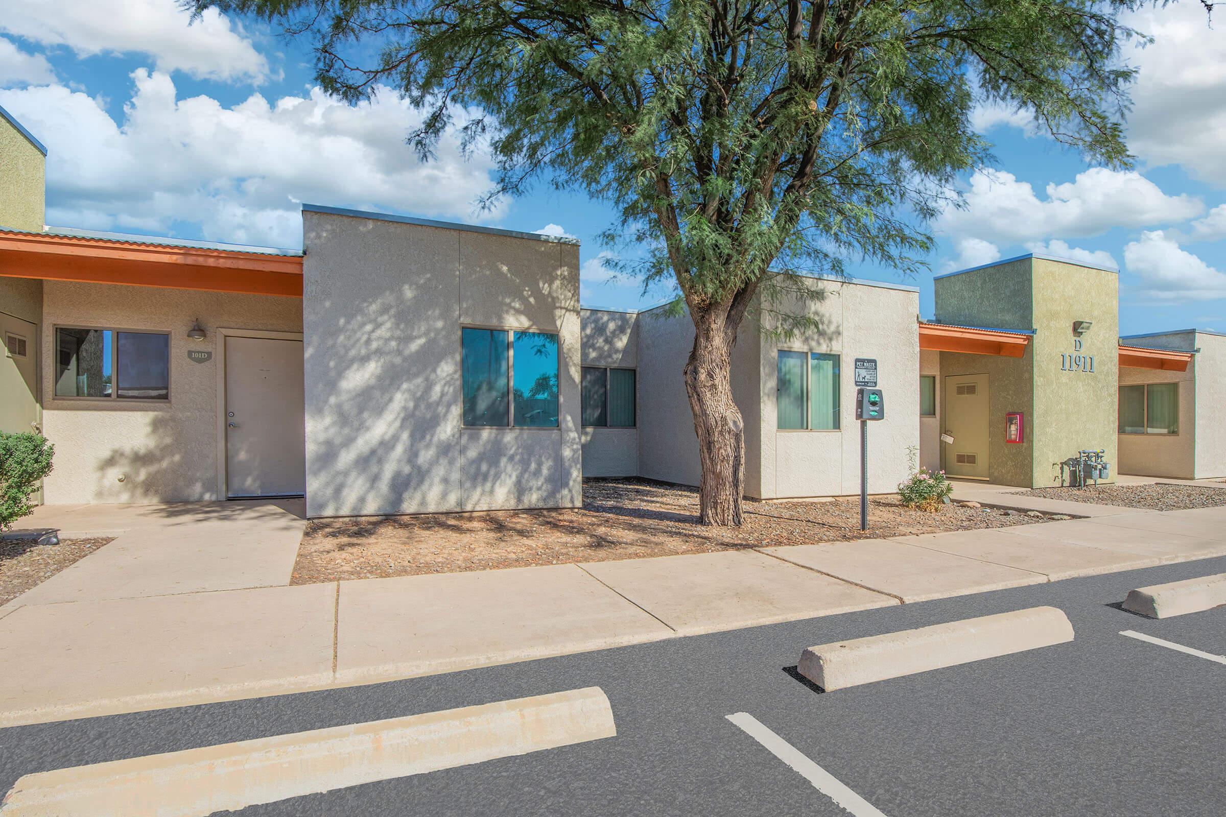 Two adjacent single-story buildings with light-colored walls and orange overhangs, surrounded by a landscaped area featuring a medium-sized tree. The scene includes a clear blue sky with fluffy clouds and a parking area with visible parking stops.