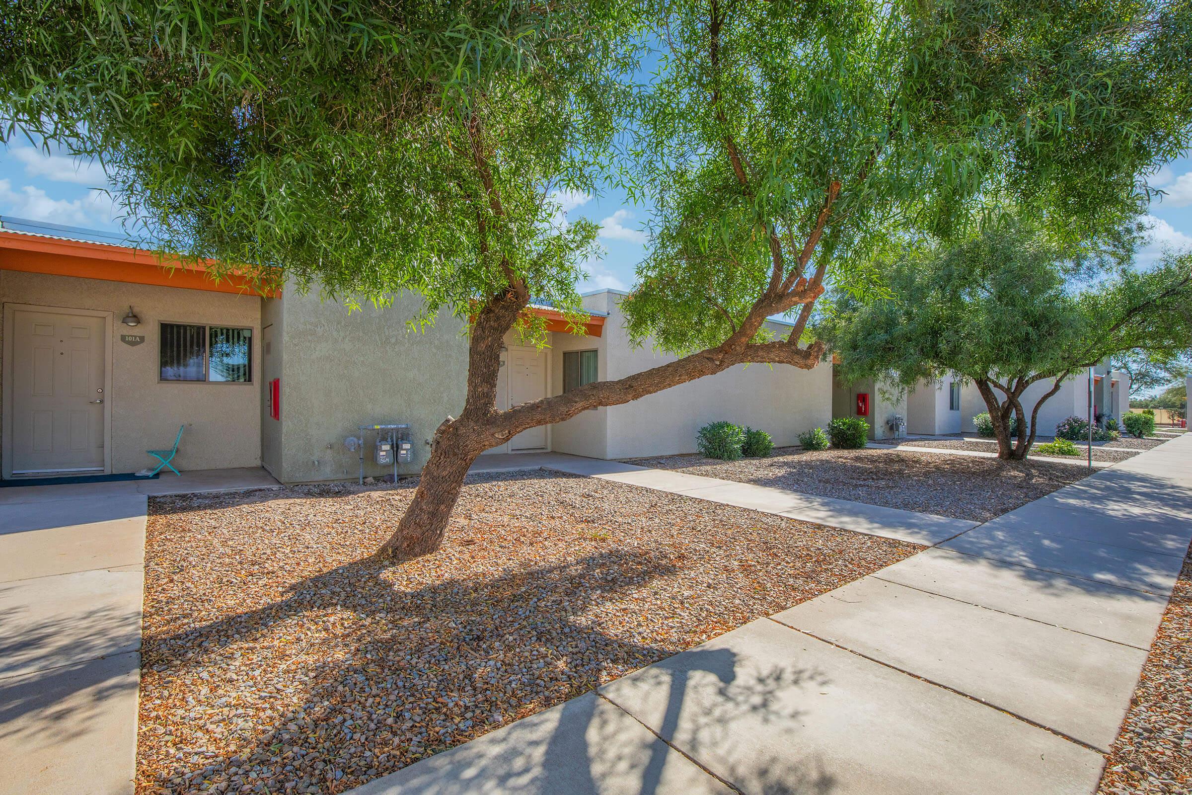A sunny exterior view of a residential complex featuring several units. Pathways lined with gravel and small shrubs lead to the entrance of the units. Trees provide shade, enhancing the inviting atmosphere of the area. The sky is clear and blue, contributing to a pleasant, serene environment.