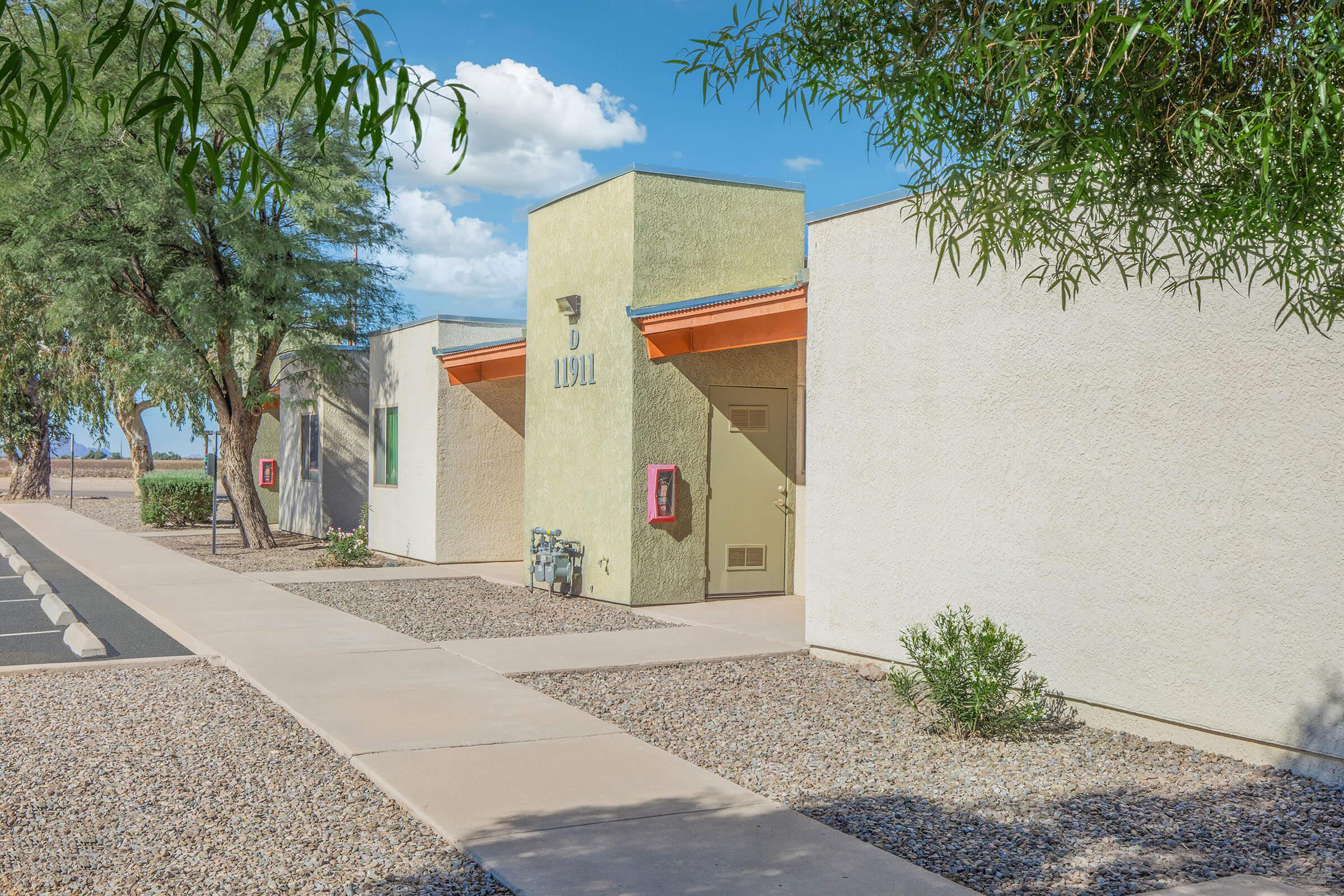 A modern, single-story building with a light-colored exterior and an orange accent on the roof. A paved walkway leads to the entrance, surrounded by landscaped gravel and a few small plants. In the background, there are trees and a clear blue sky with scattered clouds.
