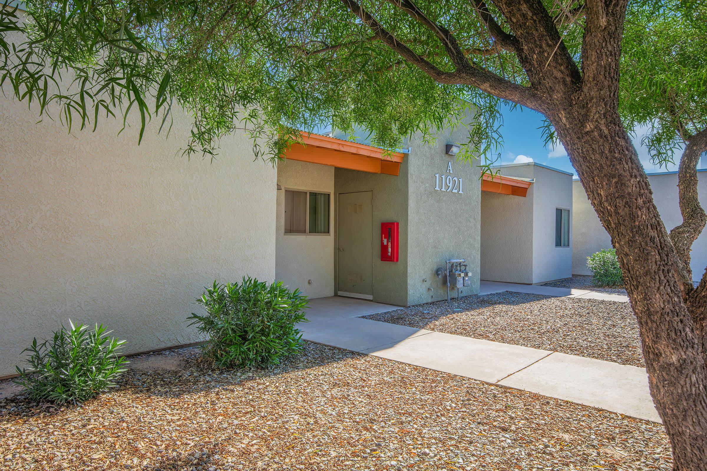 Exterior view of a beige multifamily building featuring a red fire extinguisher mounted on the wall. The entrance is framed by a large tree and shrubs, with a clear blue sky in the background. A walking path leads to the door, and gravel landscaping surrounds the base of the building.