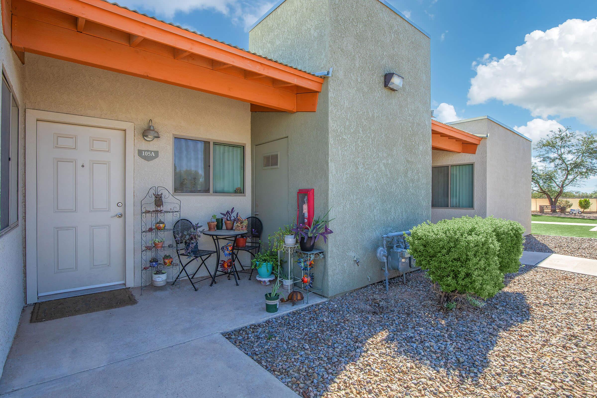 A cozy exterior view of a residential building with a well-maintained entrance featuring a small seating area with chairs and potted plants. The building has a light green exterior with an orange overhang, and a pathway leads up to the door. In the background, there are blue skies and a green lawn.
