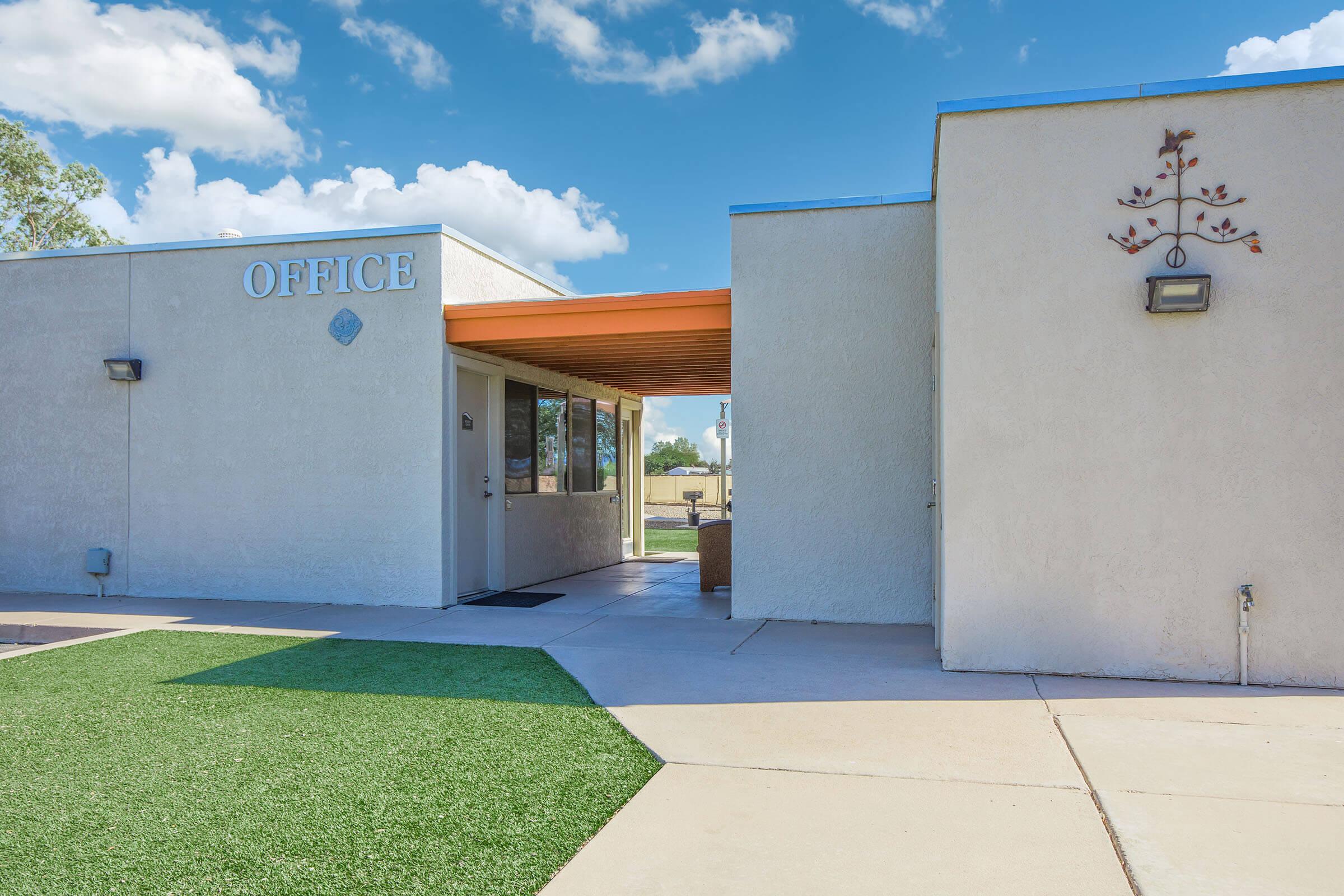 Exterior view of a building with the word "OFFICE" prominently displayed. The entrance features a clear door and an overhang. The landscape includes a small patch of green grass and paved walkways, under a bright blue sky with fluffy white clouds.