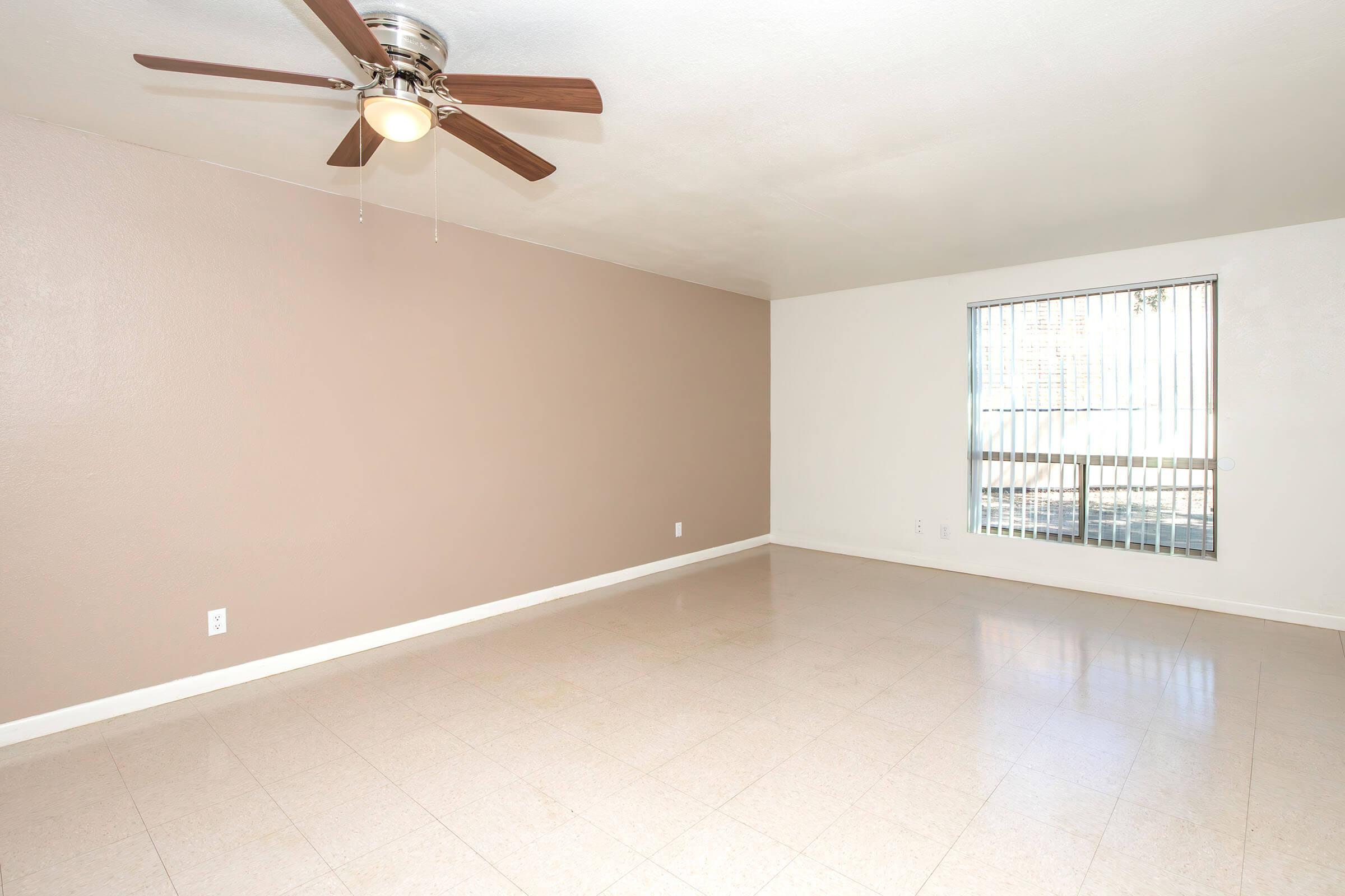 Spacious, empty living room featuring a ceiling fan, beige walls, and light-colored tile flooring. Large window with blinds allows natural light to enter. Ideal for personal decoration and furniture arrangement. Bright and airy atmosphere in a modern setting.