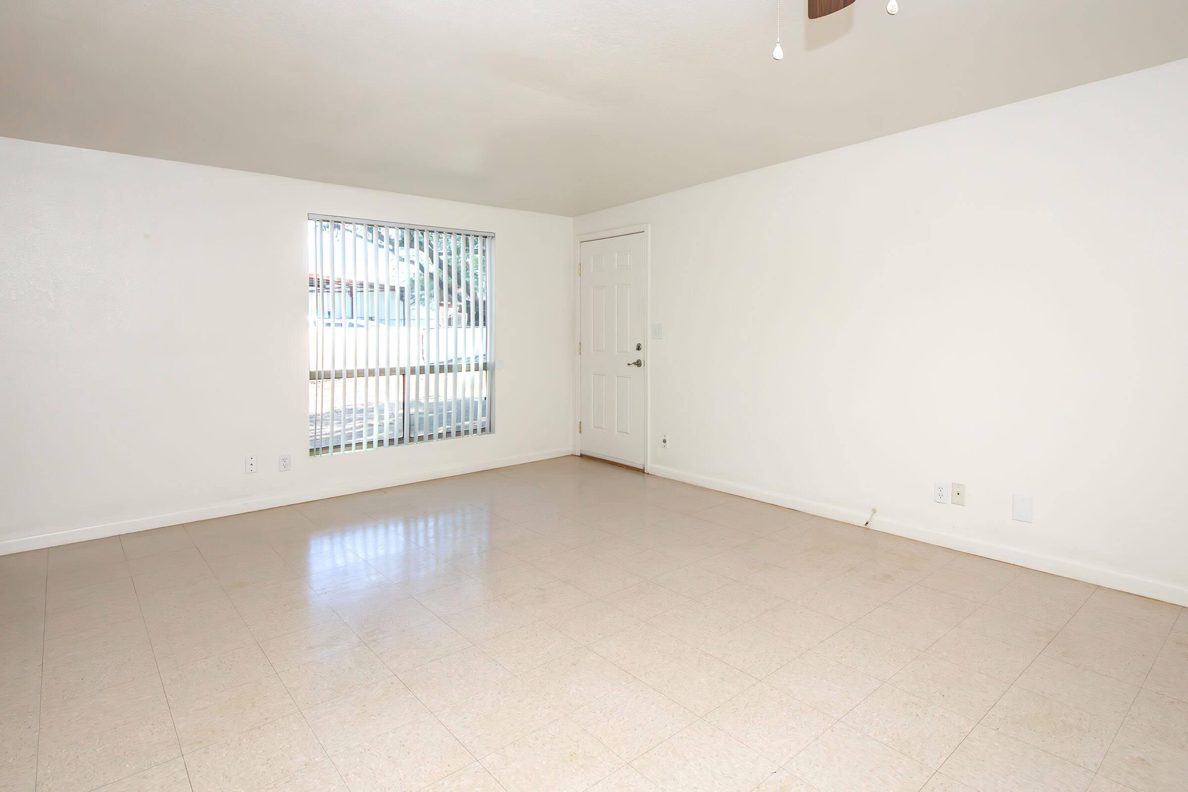 A spacious, empty room with white walls and a light-colored tiled floor. There is a large window with vertical blinds allowing natural light to enter. A door is visible on the right side of the wall. The space has a ceiling fan, contributing to a clean and simple aesthetic. Ideal for personalization or furnishing.