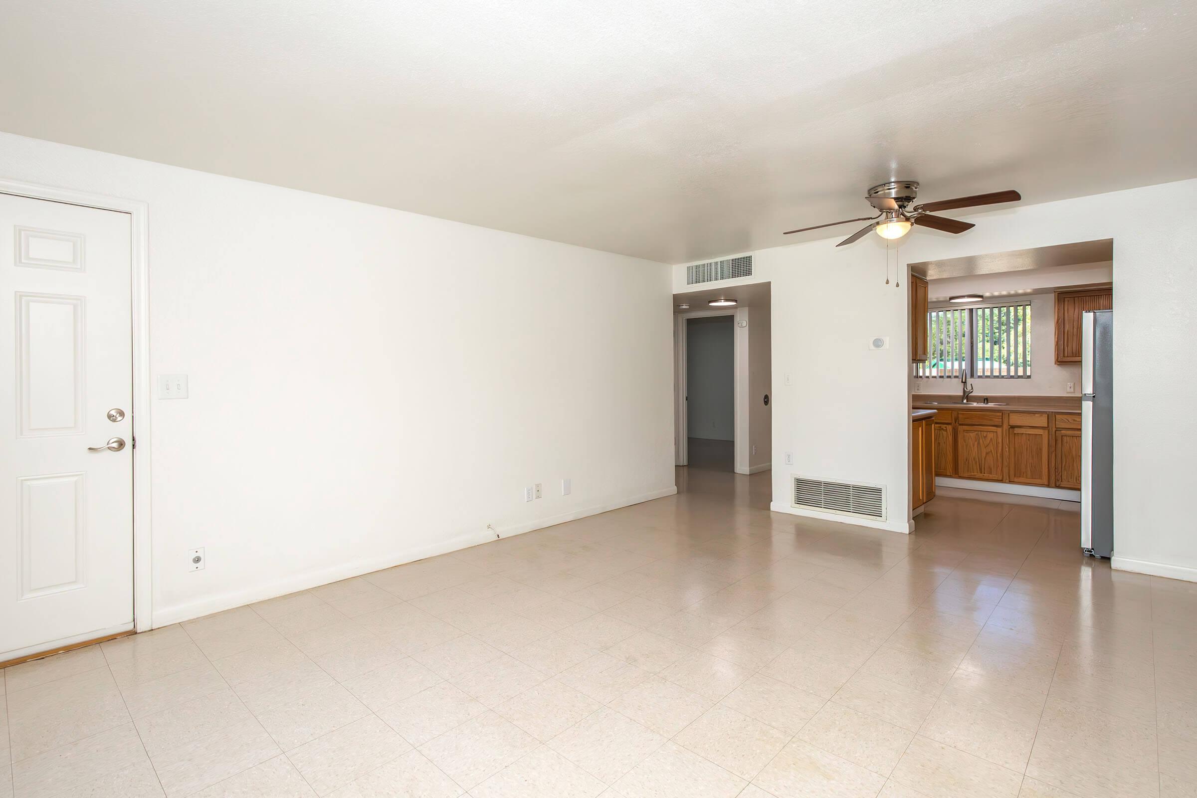 A spacious, empty living room with light-colored walls and tiled flooring. It features a ceiling fan and an open layout leading to a kitchen area with wooden cabinets. A door on the left leads outside, while a doorway on the right opens into another room. The overall atmosphere is bright and clean.